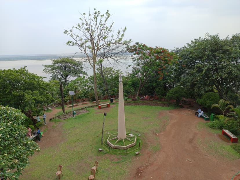 The summit of Agnigarh hill at Tezpur laid out as a public viewpoint: a stone monument column on an octagonal base set in a grassed clearing ringed by trees and benches, the spiral-stair railing at the edge, with the Brahmaputra and its sandbars visible through the trees beyond