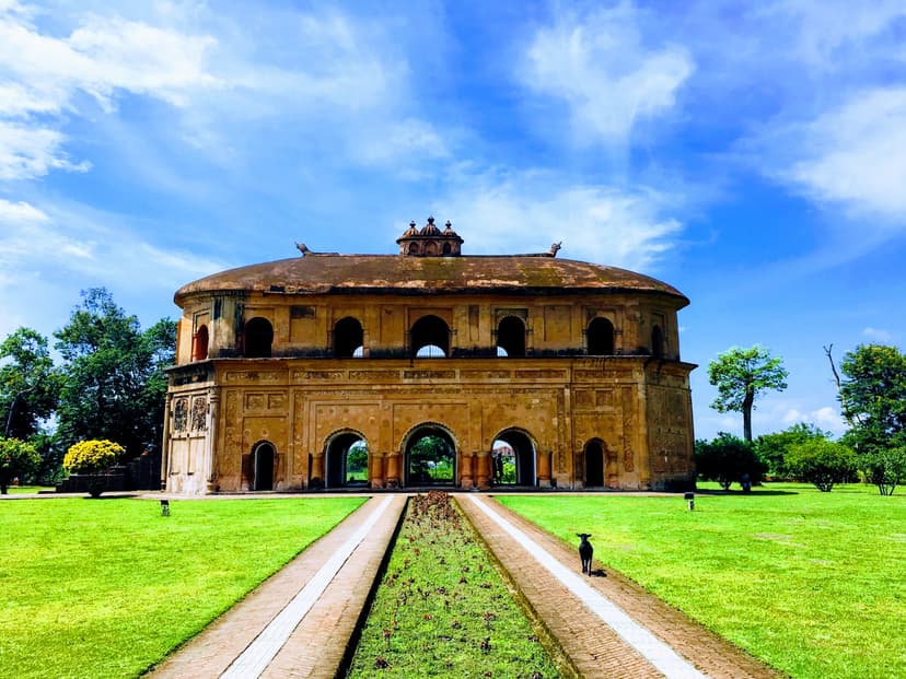 Frontal view of the Rang Ghar under a dramatic blue sky with sculptural clouds, the central garden path leading to the arched lower storey across a bright green lawn