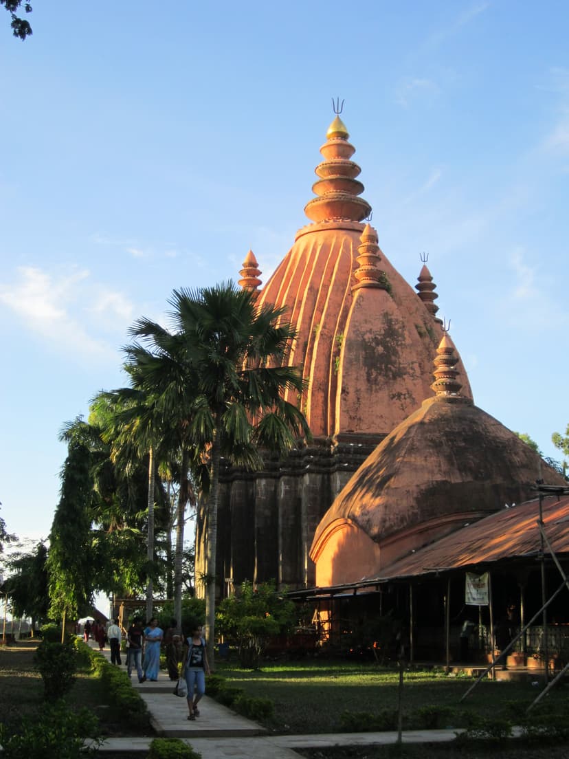 The towering red-ochre brick spire of the Sivadol at Sivasagar glowing in late-afternoon golden light against a deep blue sky, framed by a tall palm
