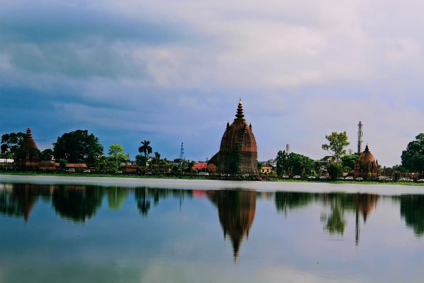 Wide view across the Sivasagar tank to the three brick temple spires of the Sivadol group on the far bank, mirrored in the still water under a moody clouded sky