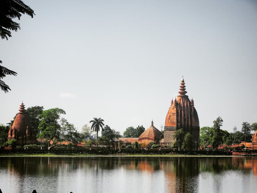 The Sivasagar tank with the Ahom temple group rising on its far bank and reflected in the water: the tall curvilinear Sivadol tower on the right, lower companion shrines to the left, the tree-lined raised bank between water and pale sky