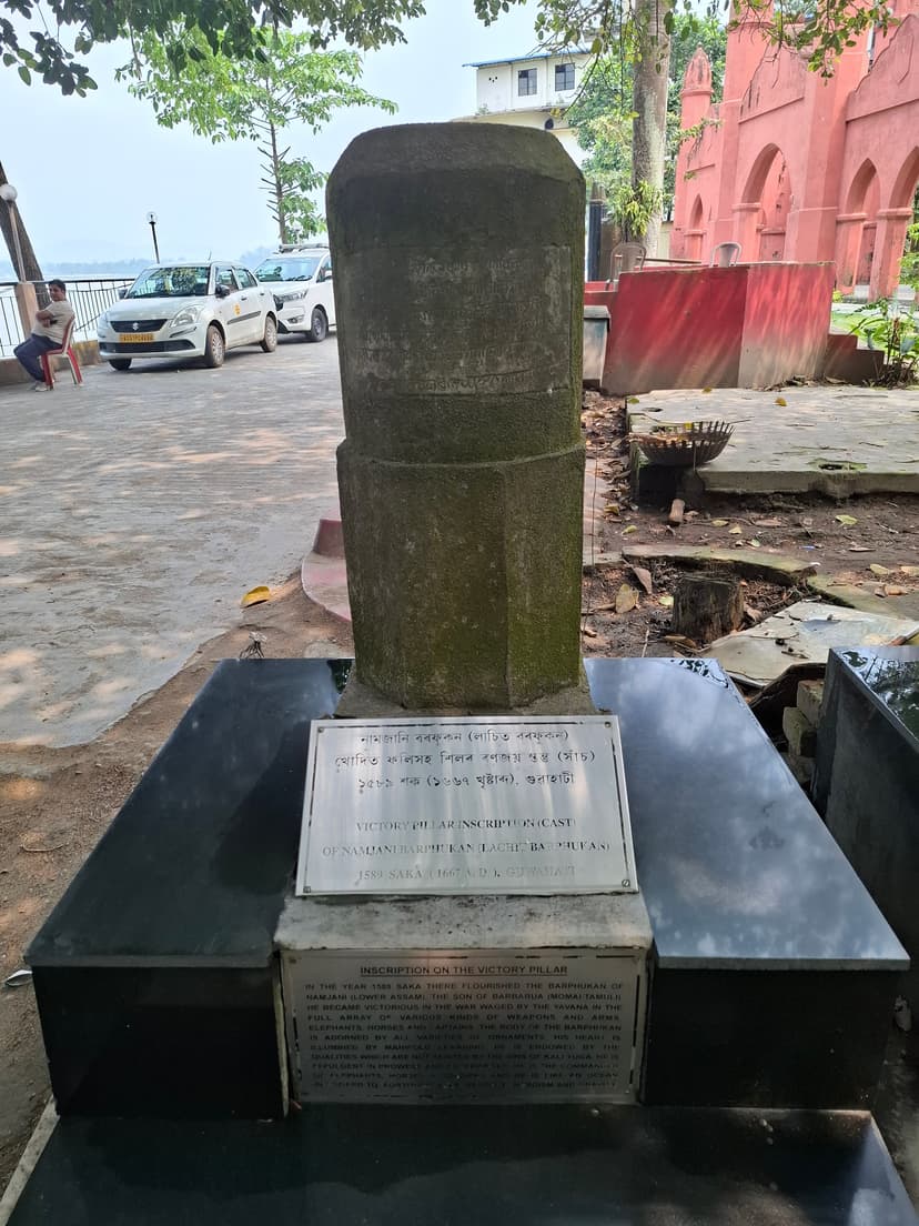 Weathered stone victory pillar of 1589 Saka on a black plinth with bilingual cast-inscription plaques at Sukreswar park, Guwahati