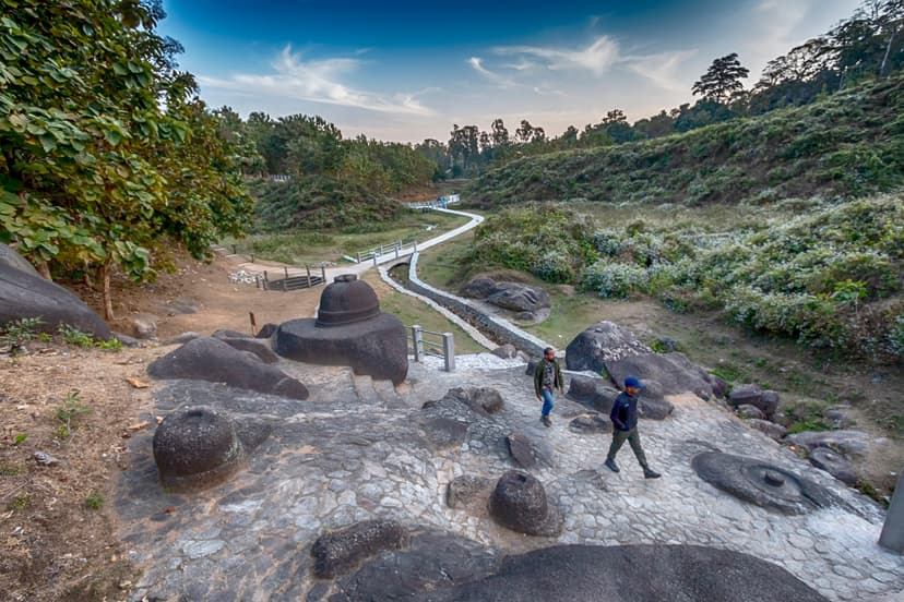 Rounded votive stupas cut from dark granite boulders on a cleared slope, a white stone path winding down into a green valley under a golden evening sky
