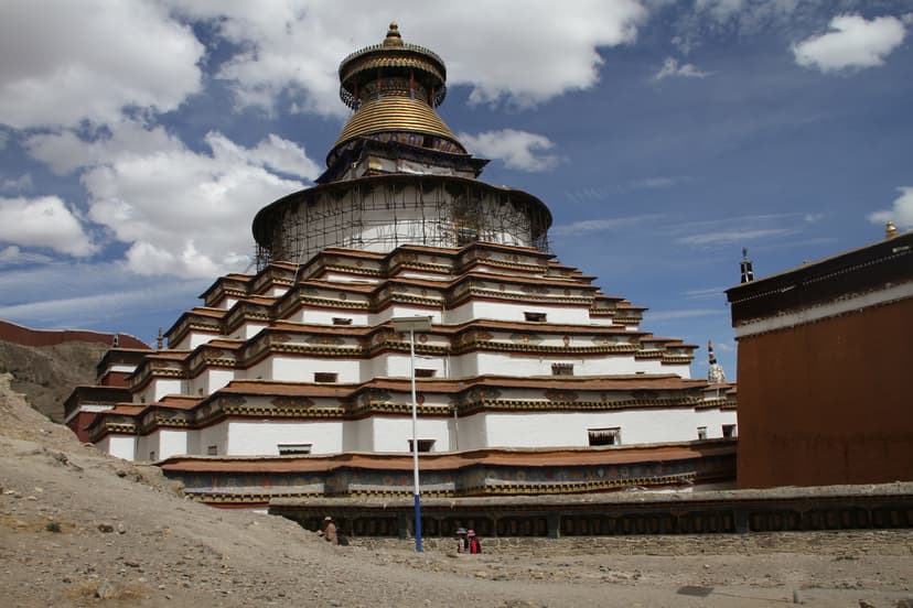 The great tiered white stupa of the Palcho Monastery at Gyantse in Tibet, its many stepped storeys banded in red and gold beneath a golden crowning dome, set against a bare high-altitude hillside and a blue sky of broken cloud