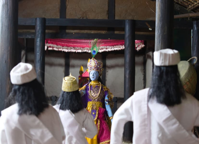 A bhaona scene: a blue-painted figure of Krishna in a jewelled peacock-feather crown and purple-and-gold robe stands beneath a red canopy in a thatched namghar hall, with white-clad gayan performers in white caps seen from behind in the foreground and a khol drum at the right