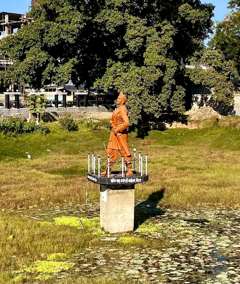 Bronze-coloured statue of a robed standing figure on a plinth set in a tank, with trees behind