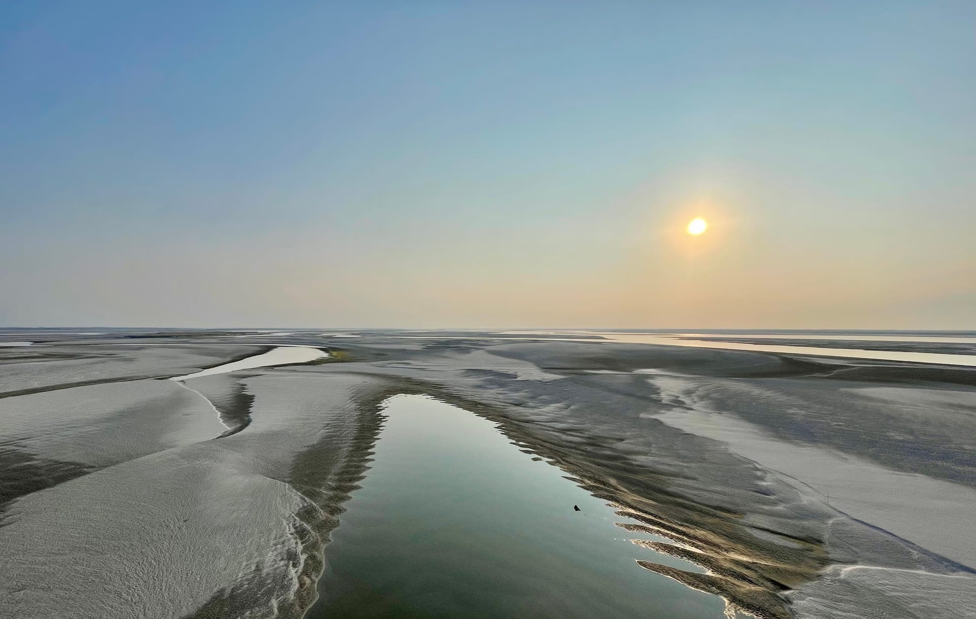 Wide view of the Brahmaputra at low water: pale sandbars and shallow channels divide the broad riverbed under a hazy sky, the low sun bright in the upper right