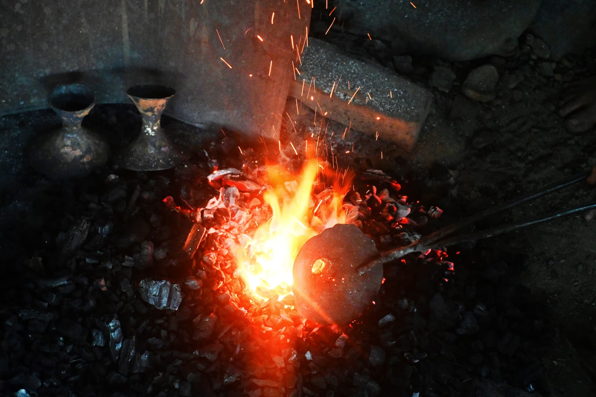 A rounded lump of bell-metal held on an iron rod glows in a bed of burning charcoal, sparks rising; two finished bell-metal vessels sit in the dark at the back of the forge