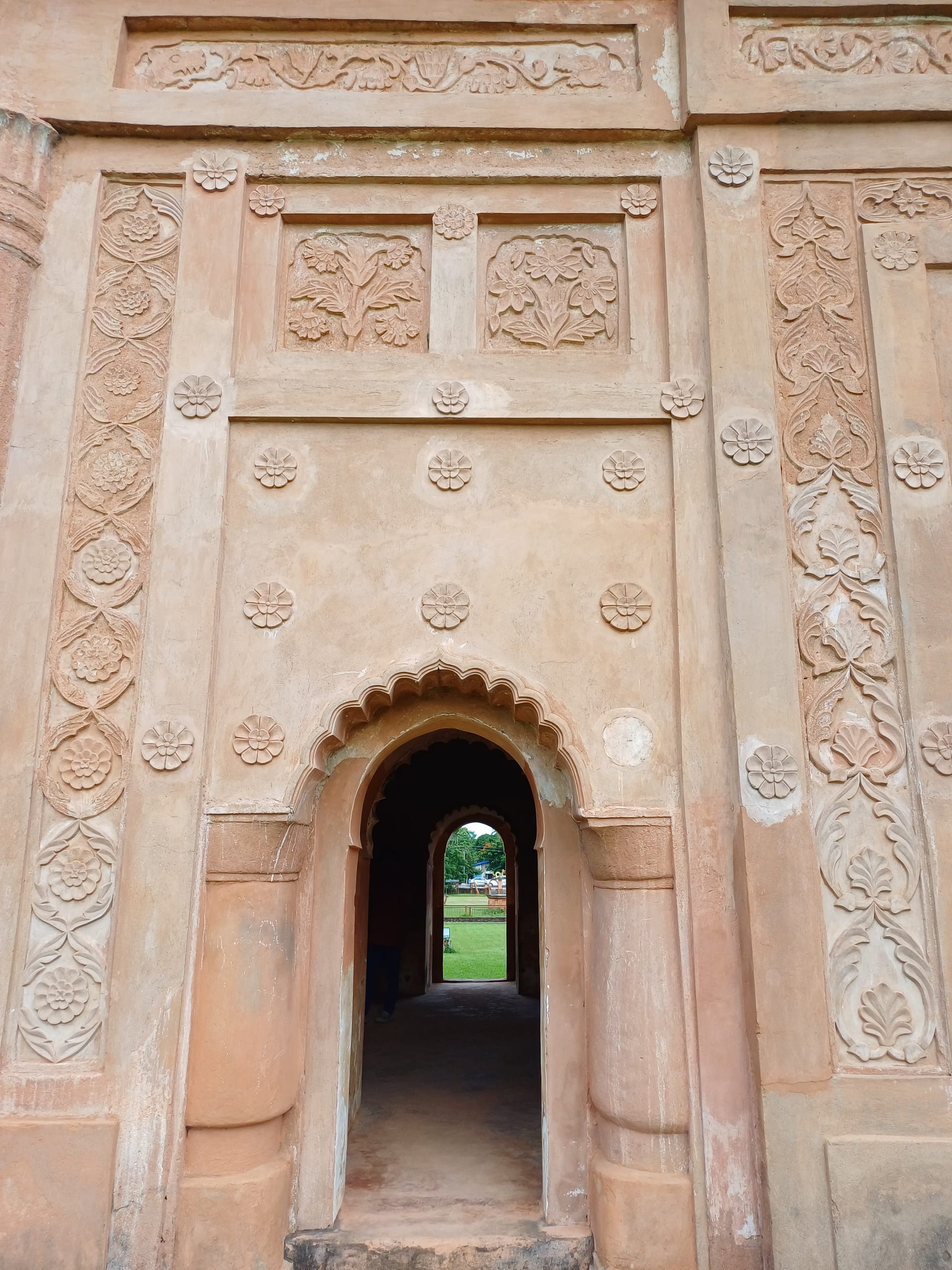 Close view of the Rang Ghar's brick wall at Sivasagar, covered in moulded terracotta ornament: bands of rosettes, framed floral relief panels, and a scalloped arched niche opening to greenery beyond