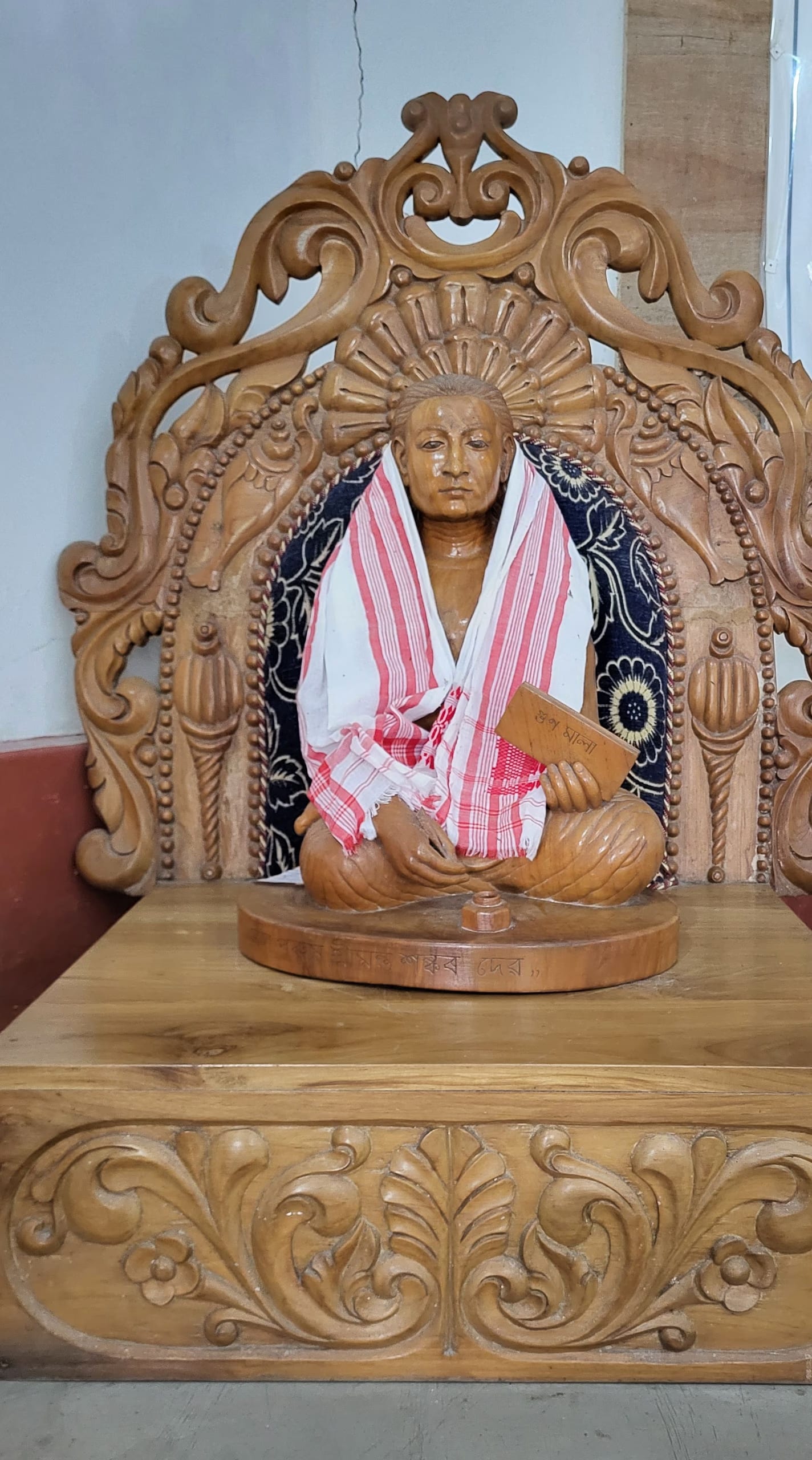 Carved wooden statue of Srimanta Sankardev seated cross-legged within an ornately carved wooden throne with a sunburst halo behind his head, draped in a red-and-white gamosa over a dark floral shawl and holding a small manuscript, on an inscribed carved pedestal