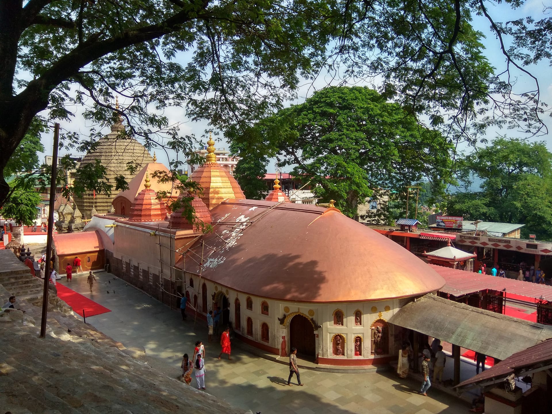 The Kamakhya temple complex seen from above: the curved copper-and-terracotta roof of the front hall, the beehive shikharas with red subsidiary domes behind, stone steps descending at left, framed by green trees