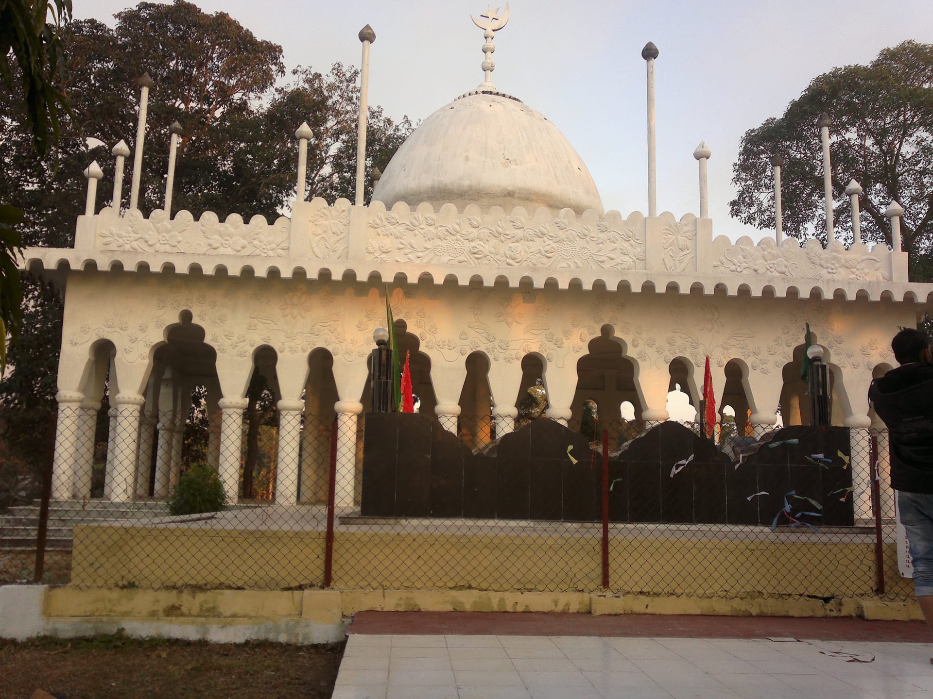 A whitewashed domed shrine with a crescent finial atop its central dome, fronted by a scalloped, floral-relief parapet and a row of arched openings; tall slender flagpoles rise around the roofline and a low chain-link fence with small flags encloses the dark-tiled platform before it.