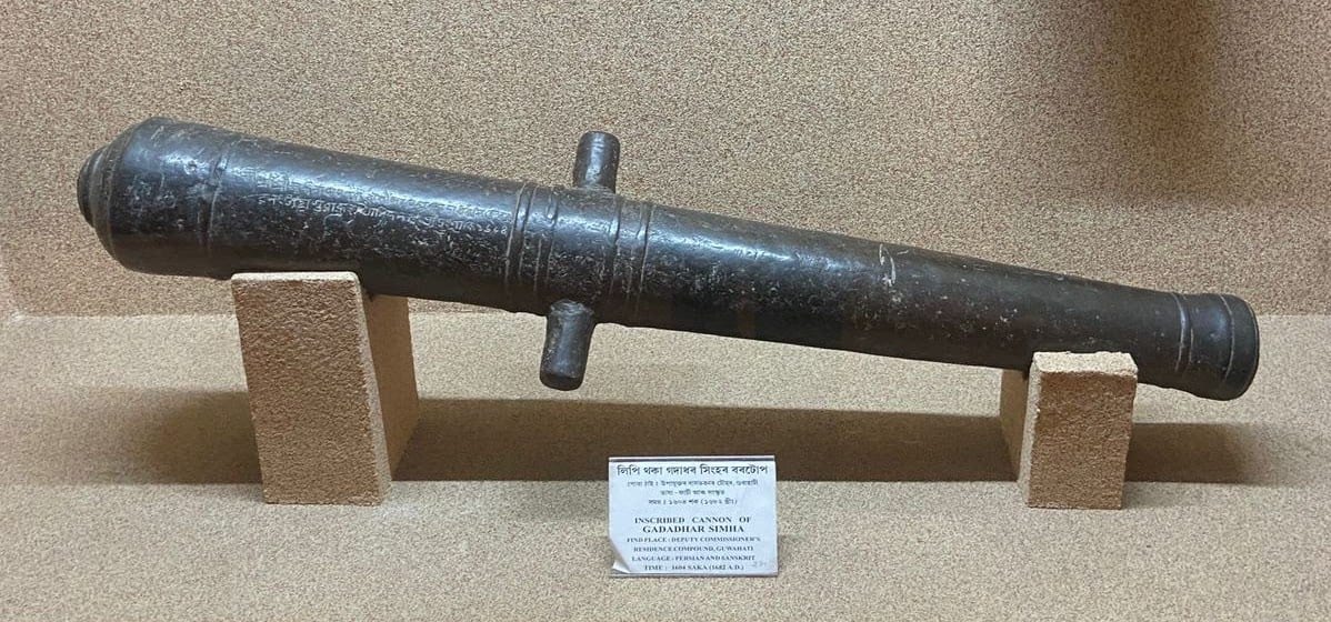 An old iron cannon resting on two brick supports in a museum display, its body bearing an Assamese inscription, with a label reading inscribed cannon of Gadadhar Simha