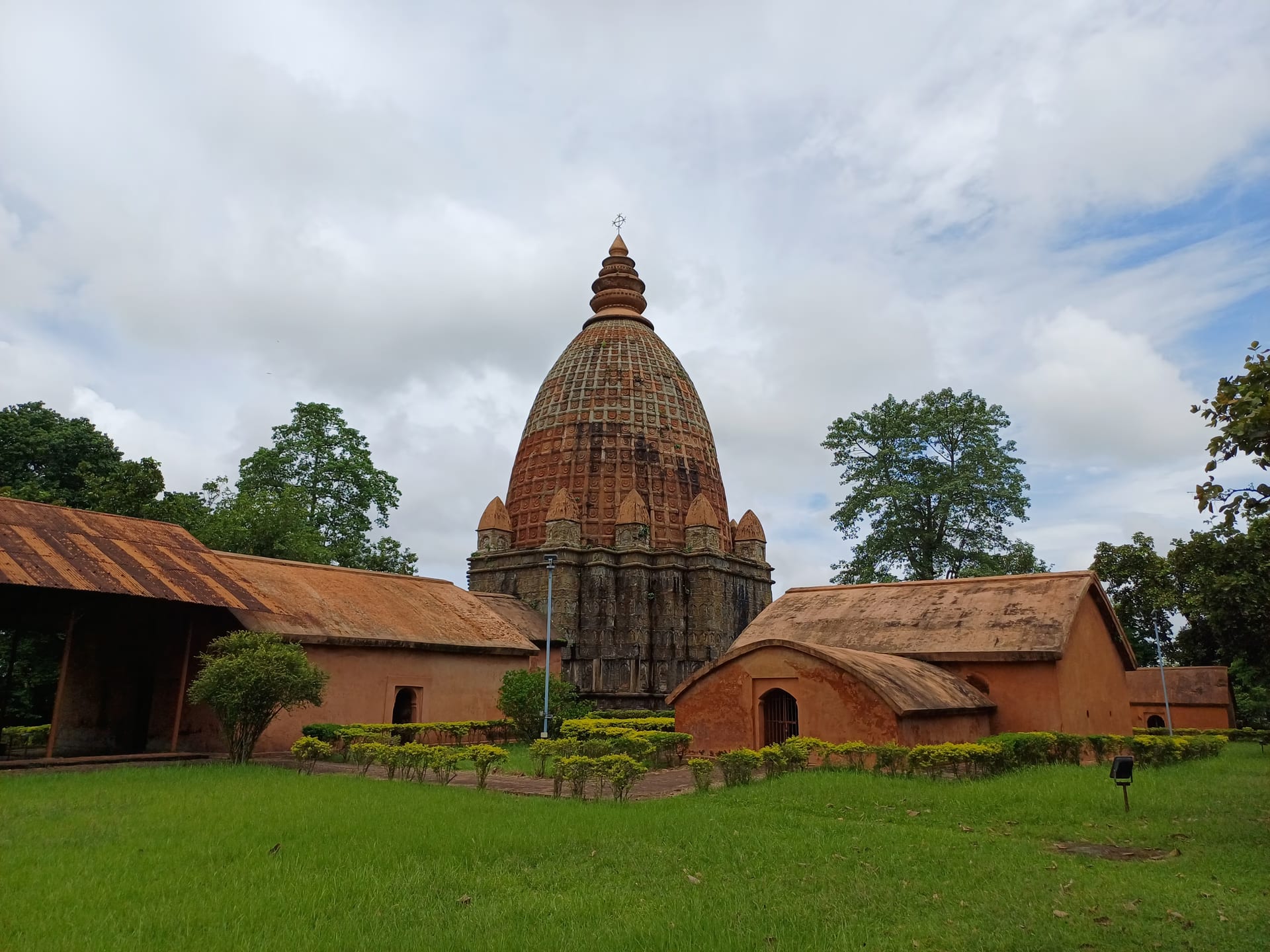 A tall brick temple with a ribbed, curvilinear spire rising over lower red-brick outbuildings with curved roofs, set behind a mown lawn and hedge under a cloudy sky
