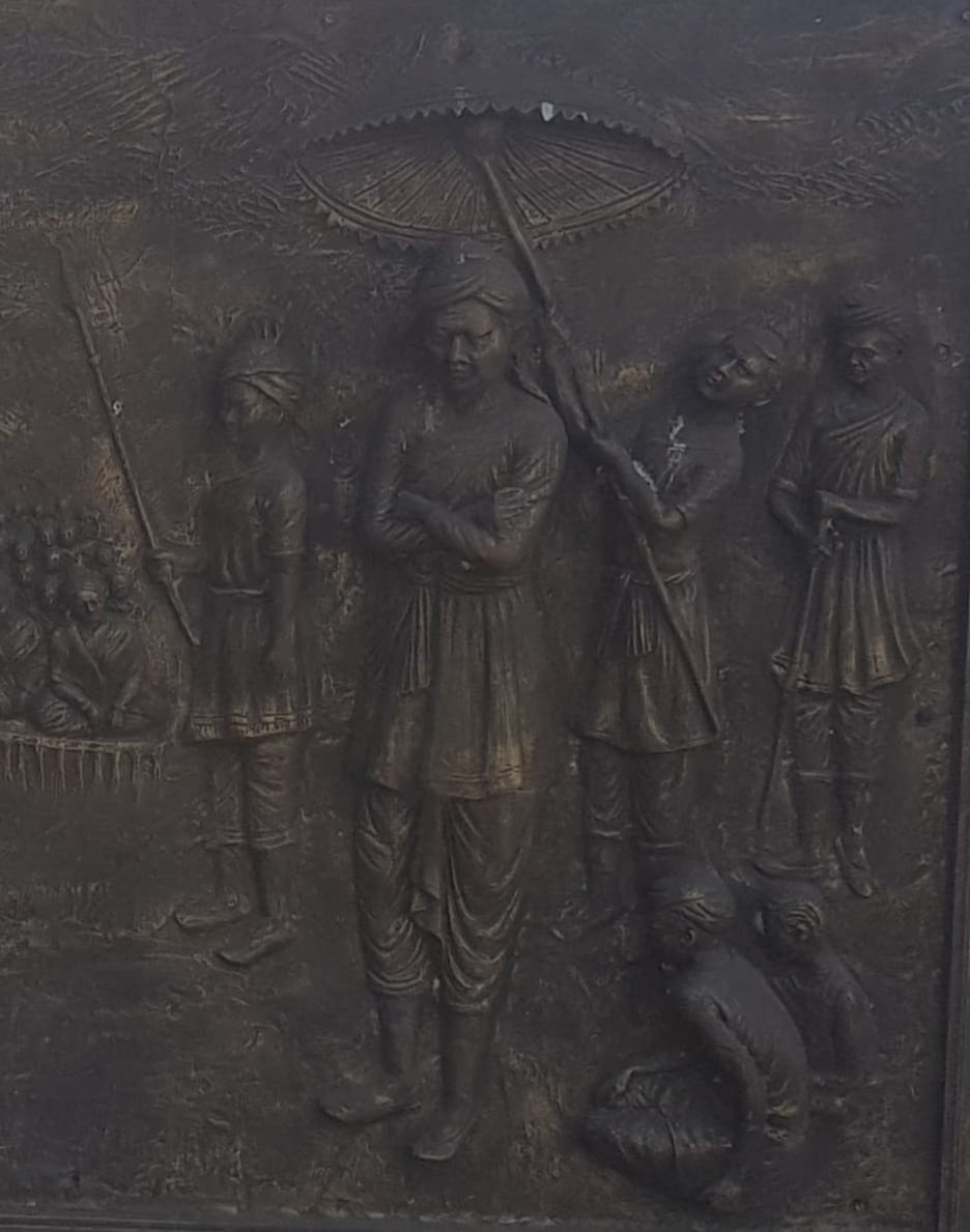 Bronze relief panel of Momai Tamuli Borbarua, the first Borbarua, standing beneath a ceremonial parasol with attendants