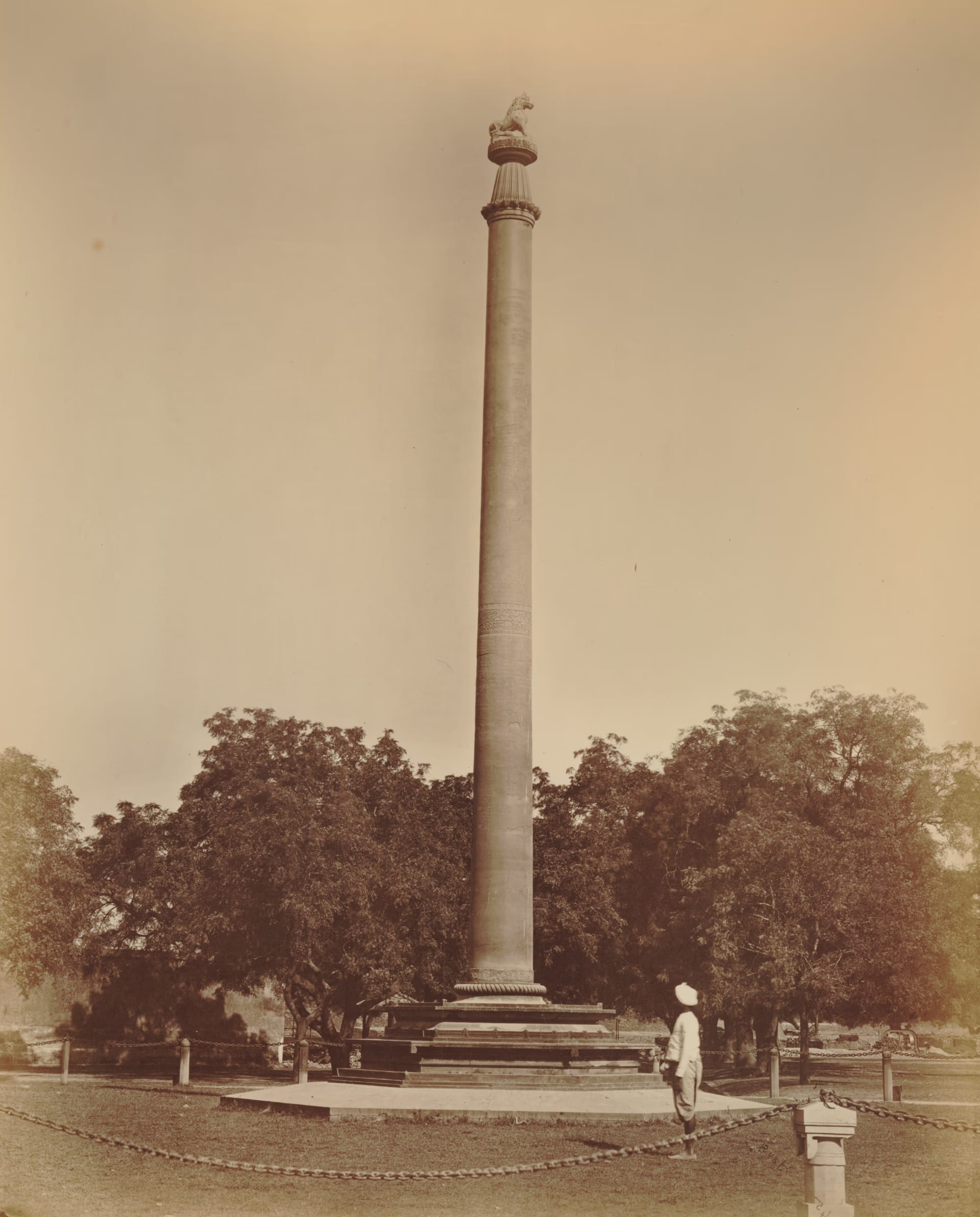 A tall polished sandstone column standing on a stepped plinth among trees, a small figure at its base for scale