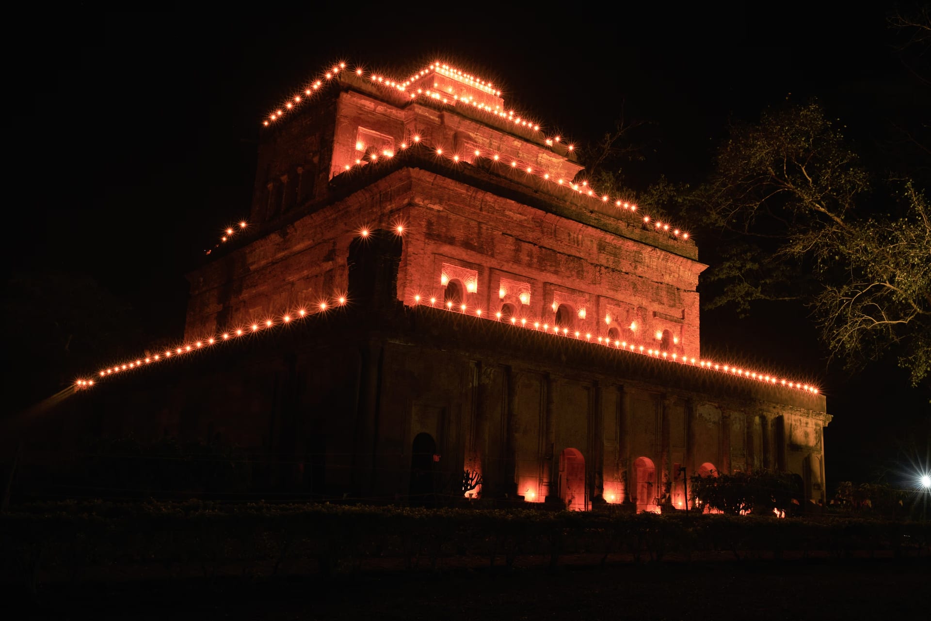 The tiered brick Kareng Ghar palace at Gargaon seen at night, its receding storeys outlined with rows of festival lamps against a black sky