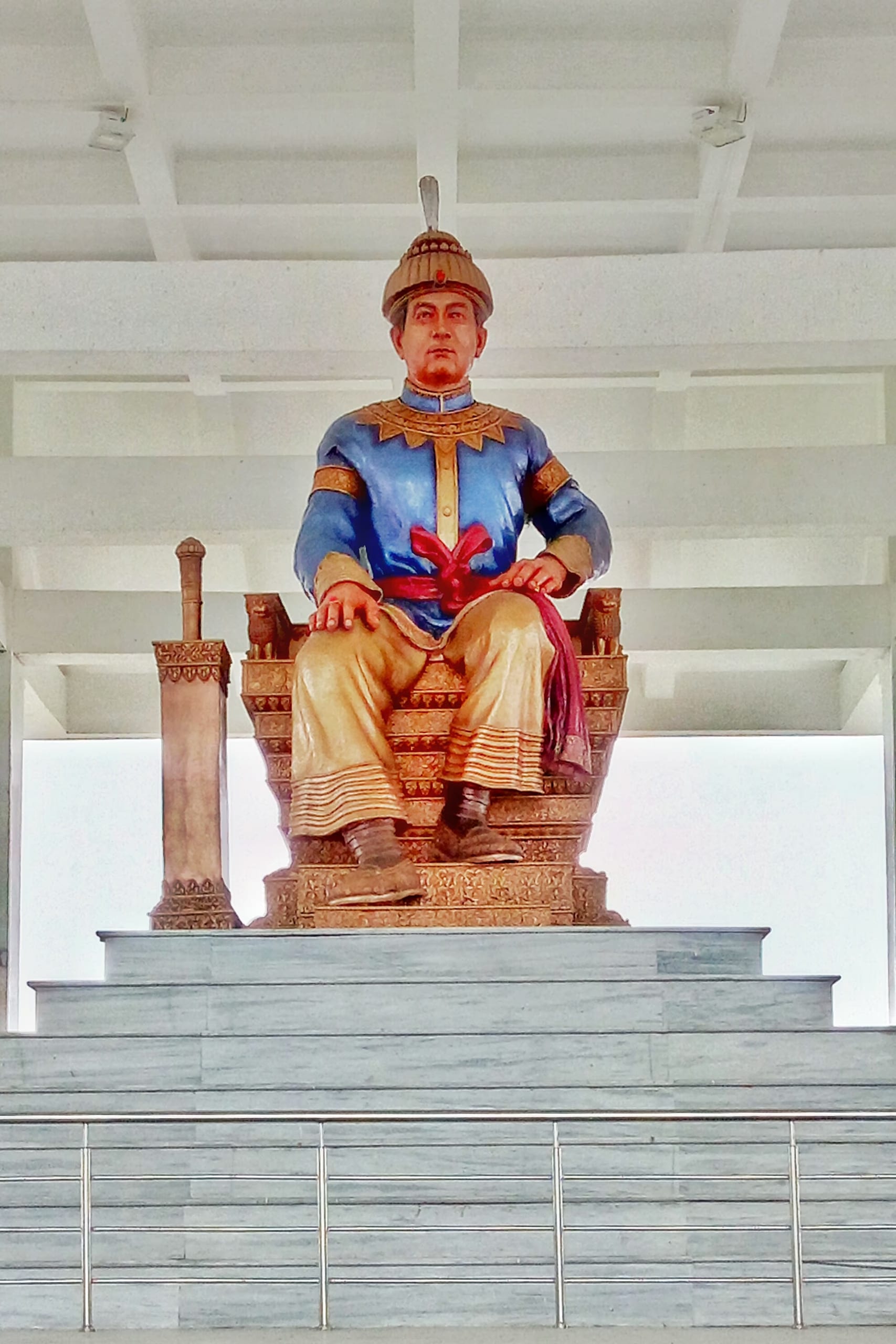 Large seated statue of Sukaphaa on a stepped marble plinth, in a blue tunic and golden dhoti, one hand resting on his knee
