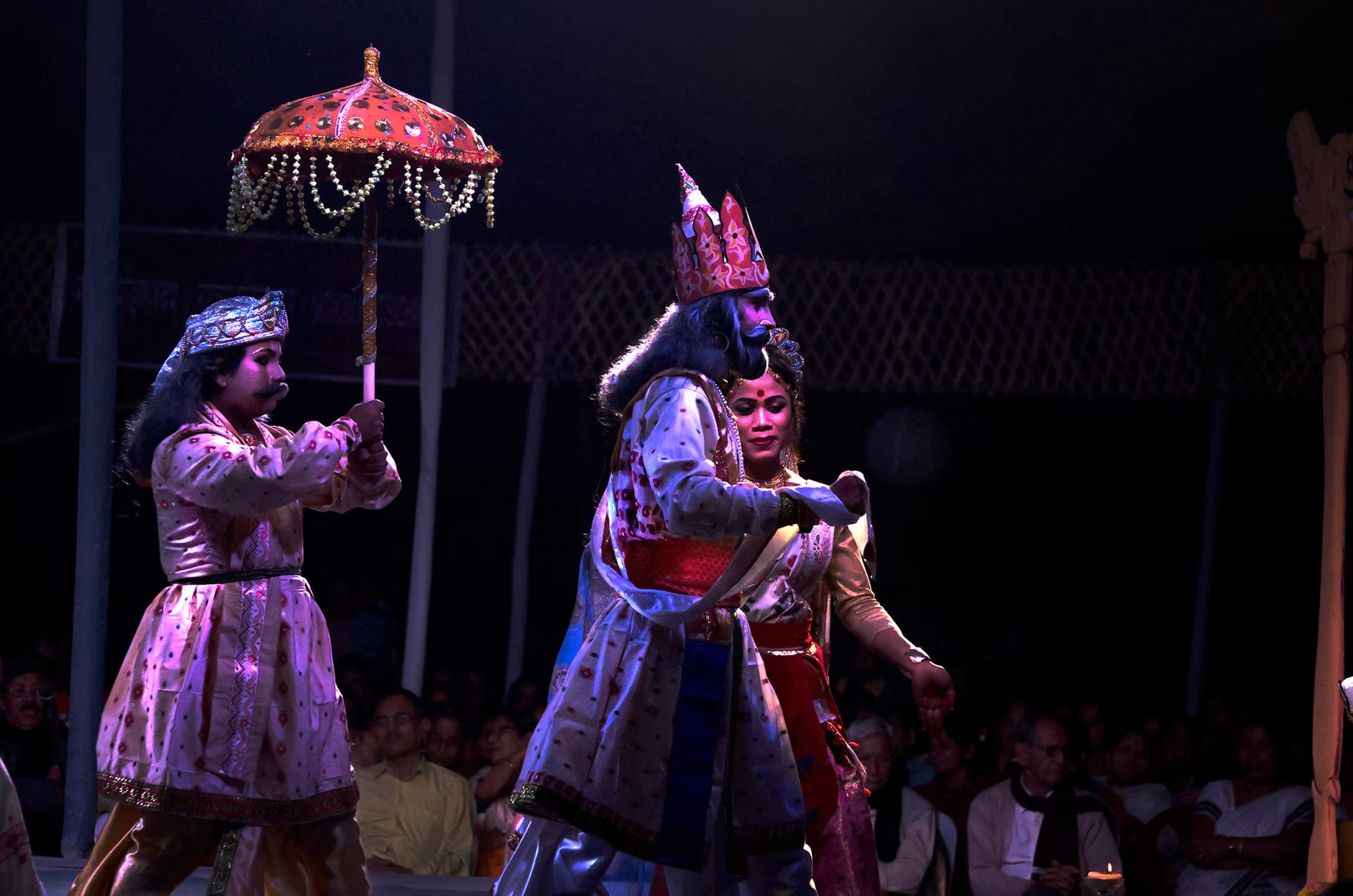 Costumed players in a night bhaona, a crowned figure under a decorated umbrella, the audience seated on the floor