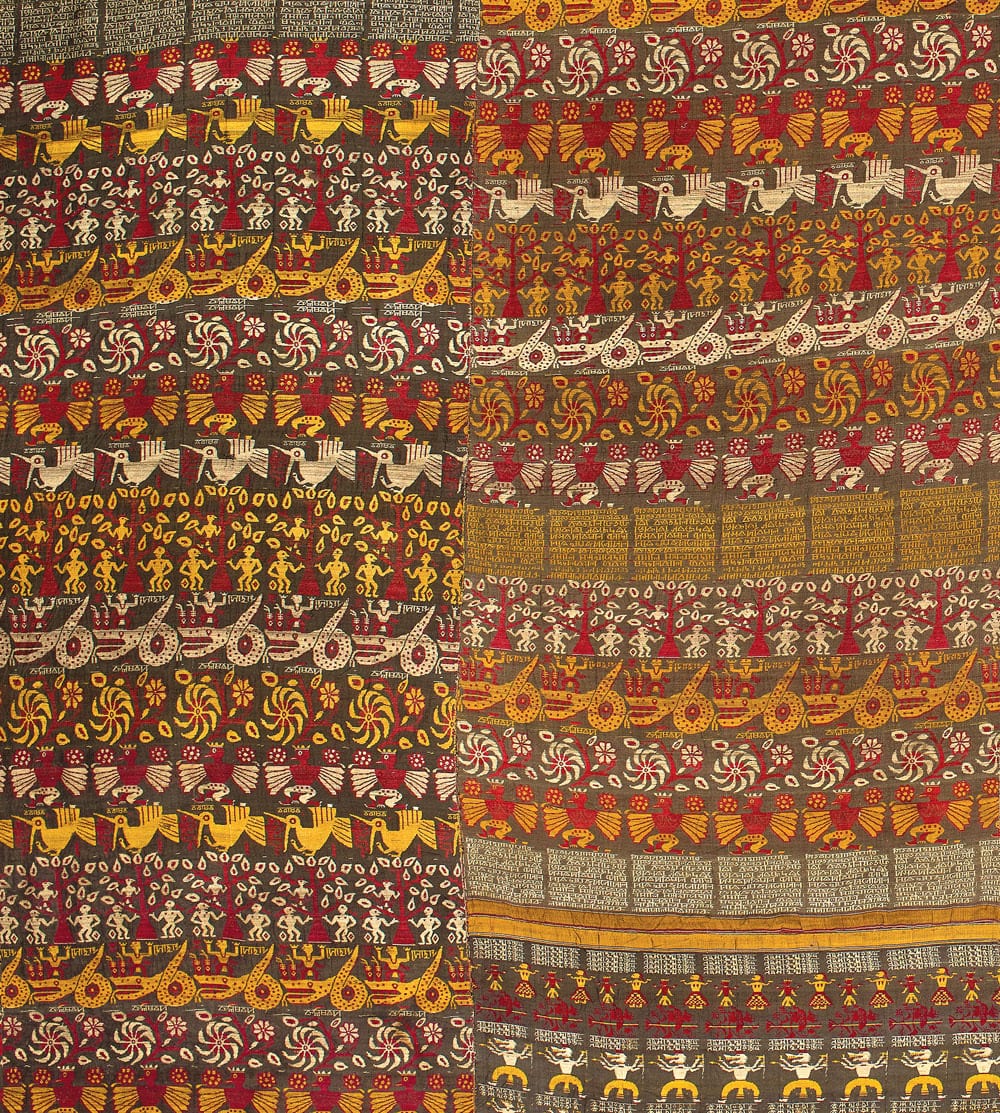 A detail of the Vrindavani Vastra silk, dark brown ground worked in red, yellow and cream with horizontal registers of winged birds, crowned figures, dancing attendants, boats and pinwheel flowers, separated by bands of woven Assamese script