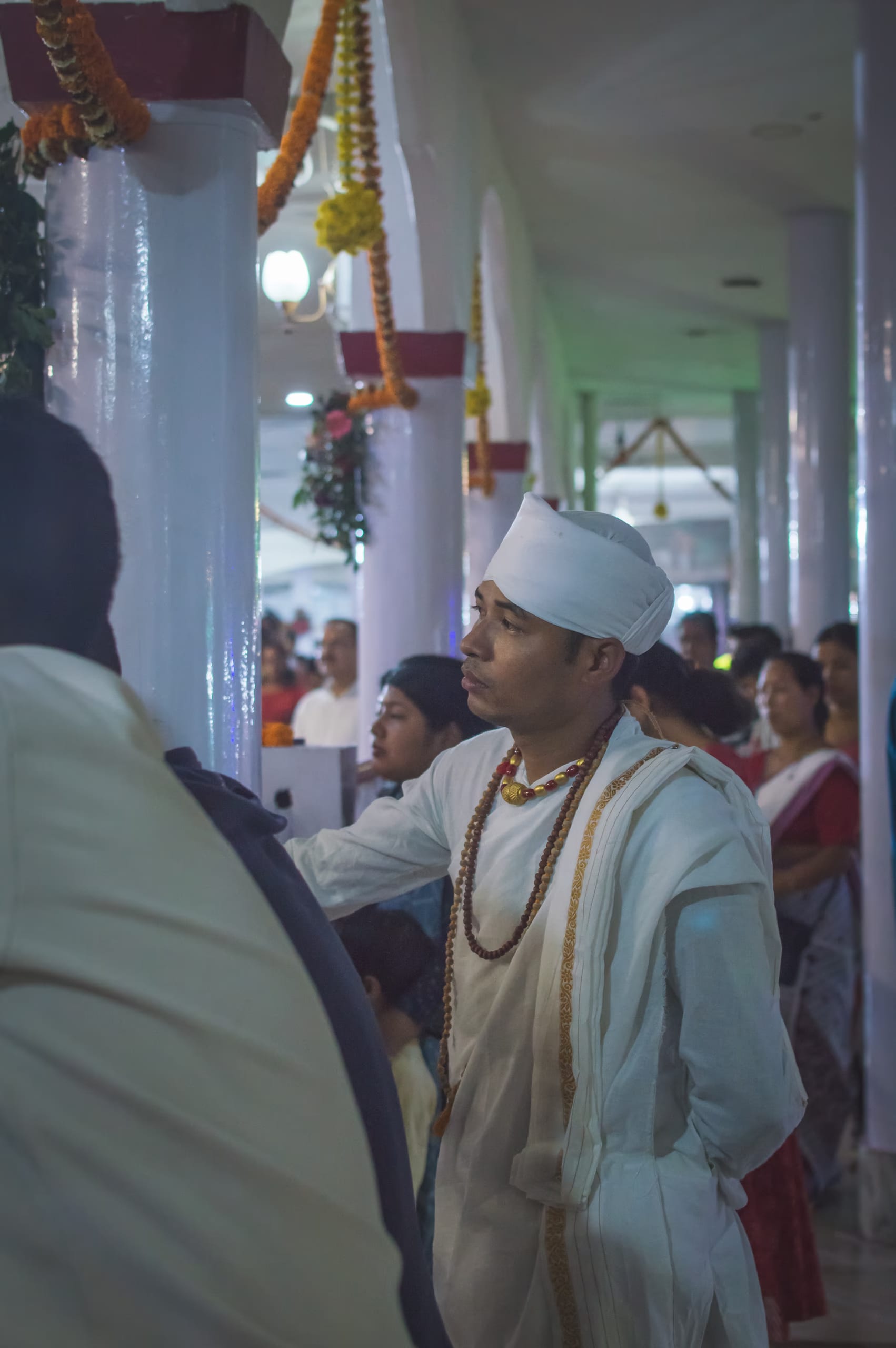 A young man in the white cloth and folded white turban of the sattra tradition, a long rudraksha rosary and a gold pendant at his neck and a gold-bordered chadar over one shoulder, standing among devotees in a garlanded pillared prayer hall