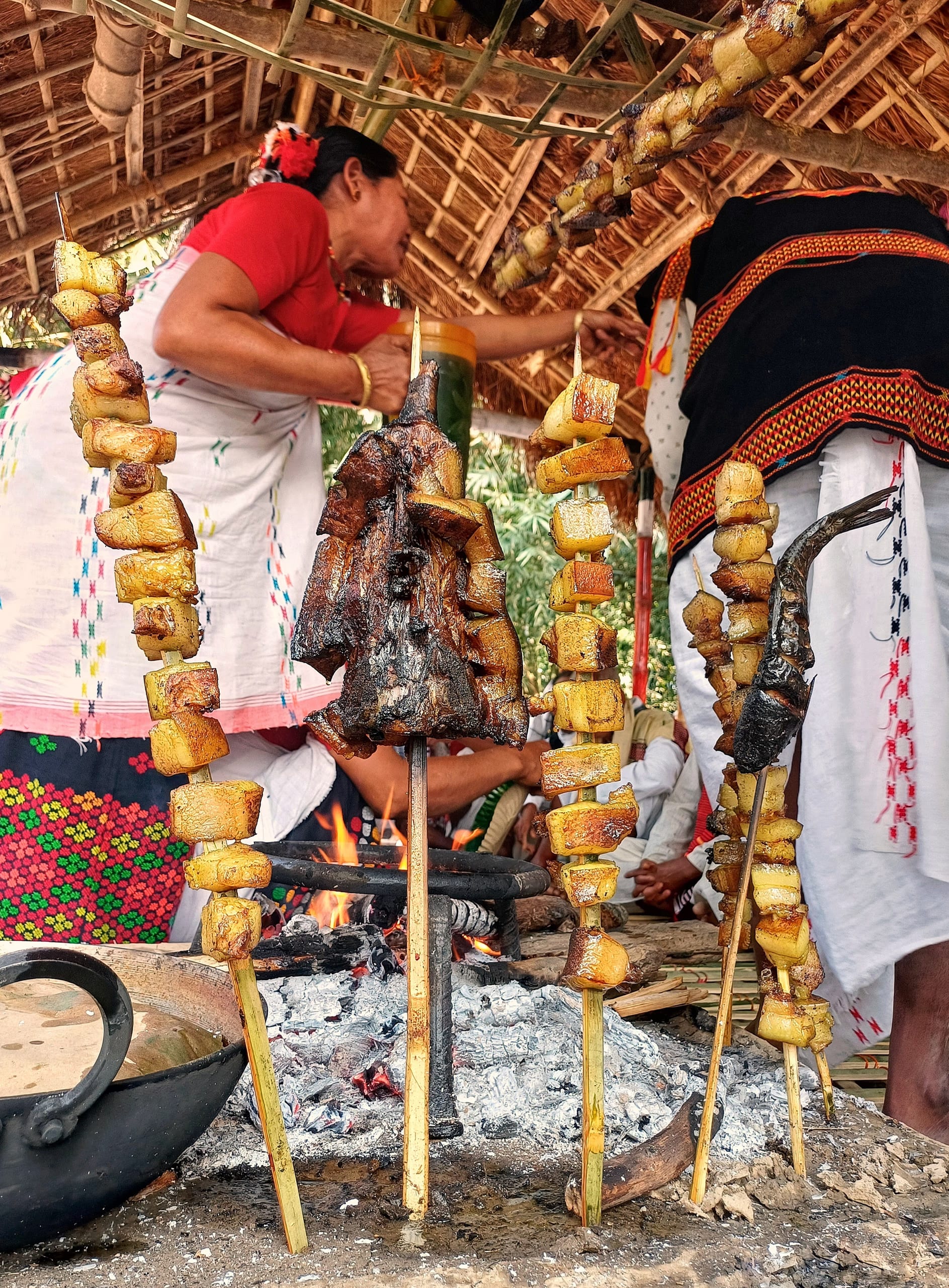 Cuts of pork and fat threaded on bamboo skewers roasting over an open fire, with women cooking behind
