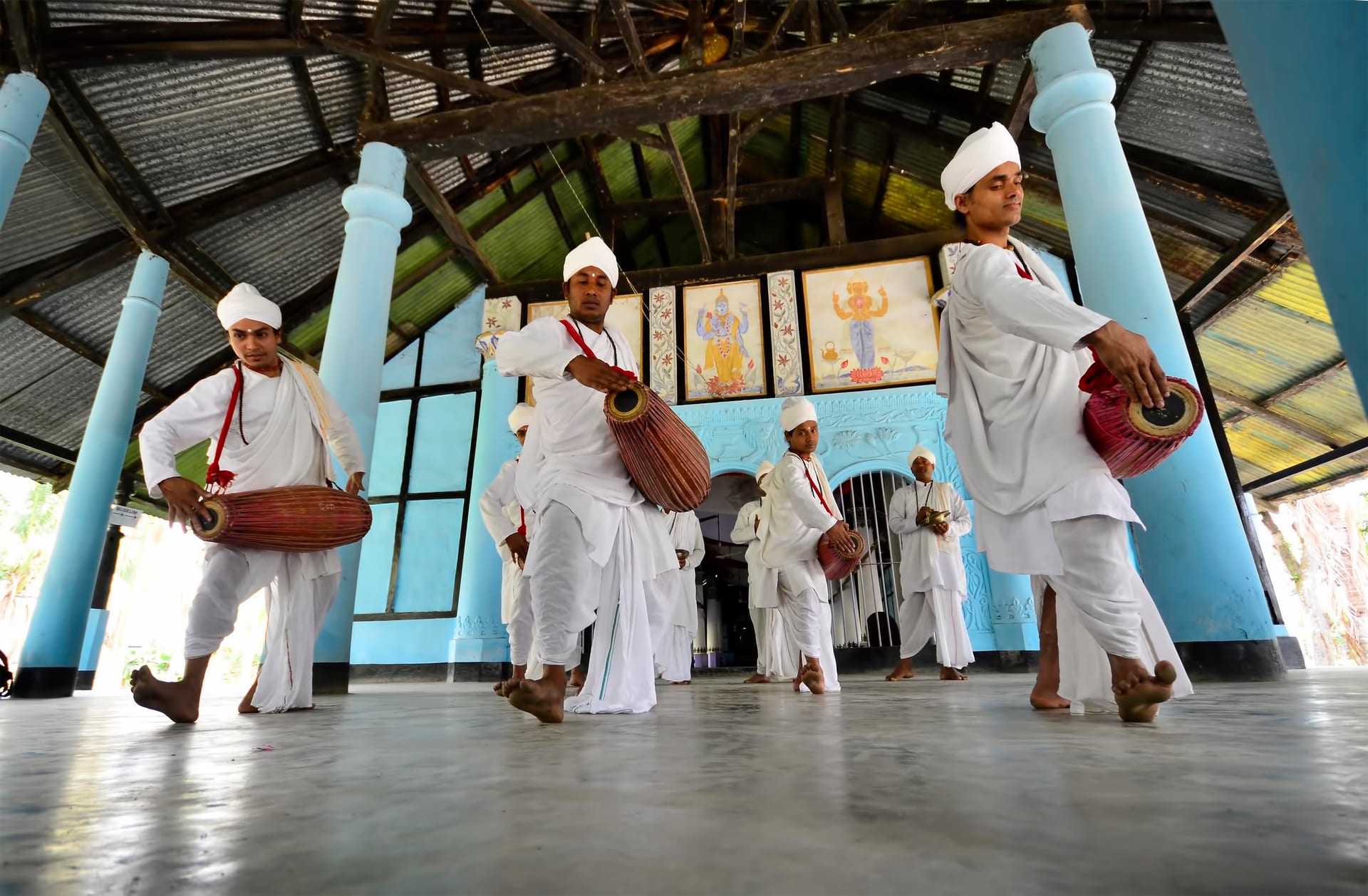 Four men in white dhotis and white turbans playing two-headed khol drums mid-stride across the floor of a sattra prayer hall in Majuli, devotional paintings of deities mounted on the blue rear wall behind them