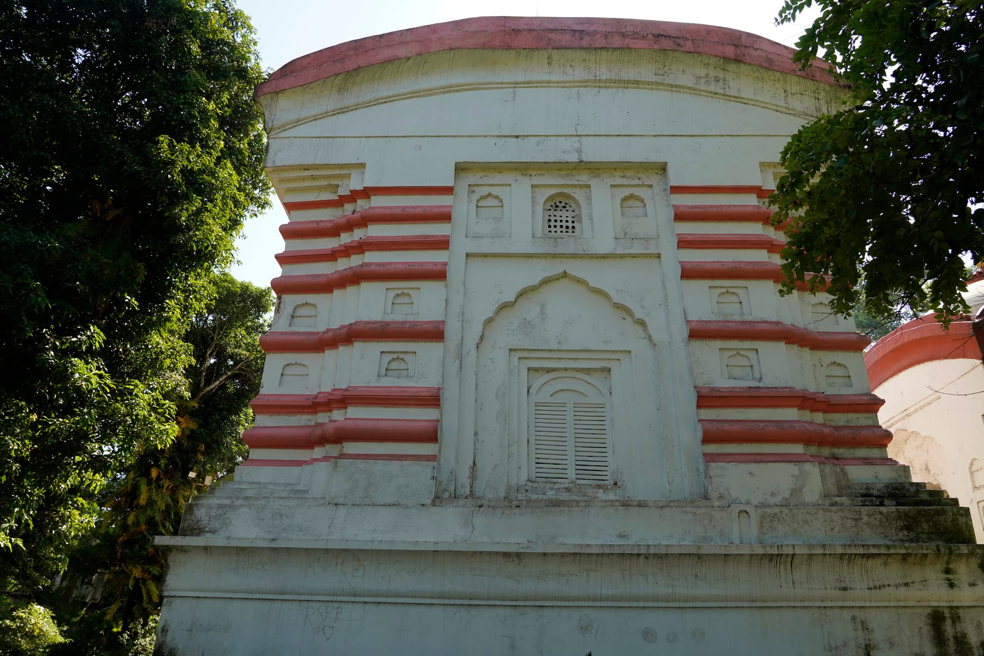 A whitewashed temple with red-painted mouldings stepping up its face in tiers, a cusped arch over the shrine door, under a curved roof, framed by trees
