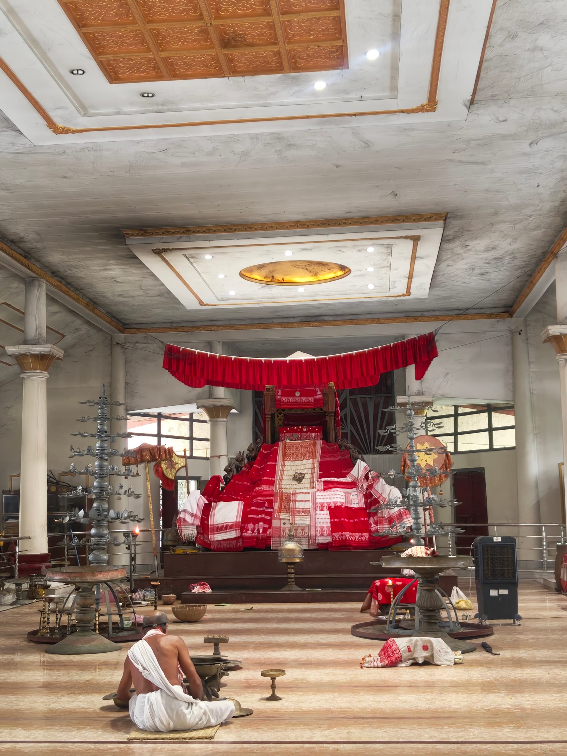 The marble-floored prayer hall of the Batadrava Than, with a red-draped throne bearing the sacred manuscript at its centre and brass ceremonial lamps to either side