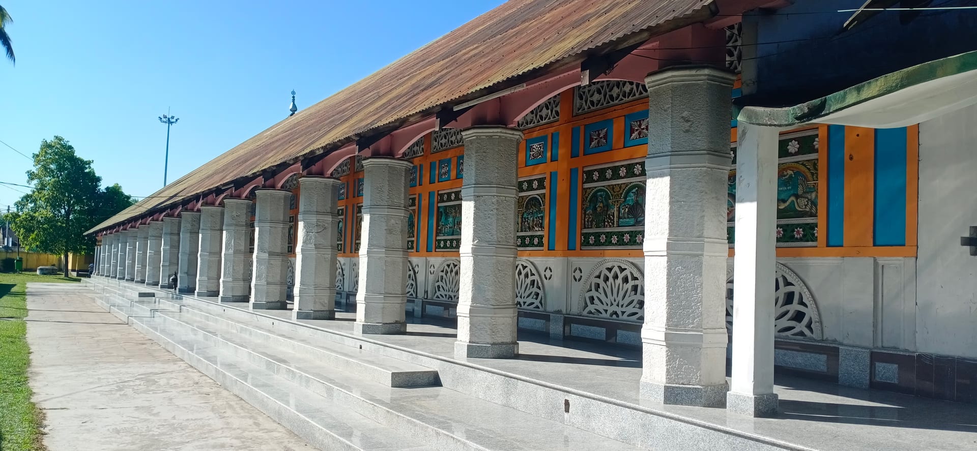 The long pillared veranda of the Barpeta Sattra kirtan-ghar in bright sunlight, a row of stout whitewashed columns on a raised plinth receding in perspective beneath a low pitched roof, the wall behind painted in orange with figural panels and pierced white lattice screens, under a clear blue sky