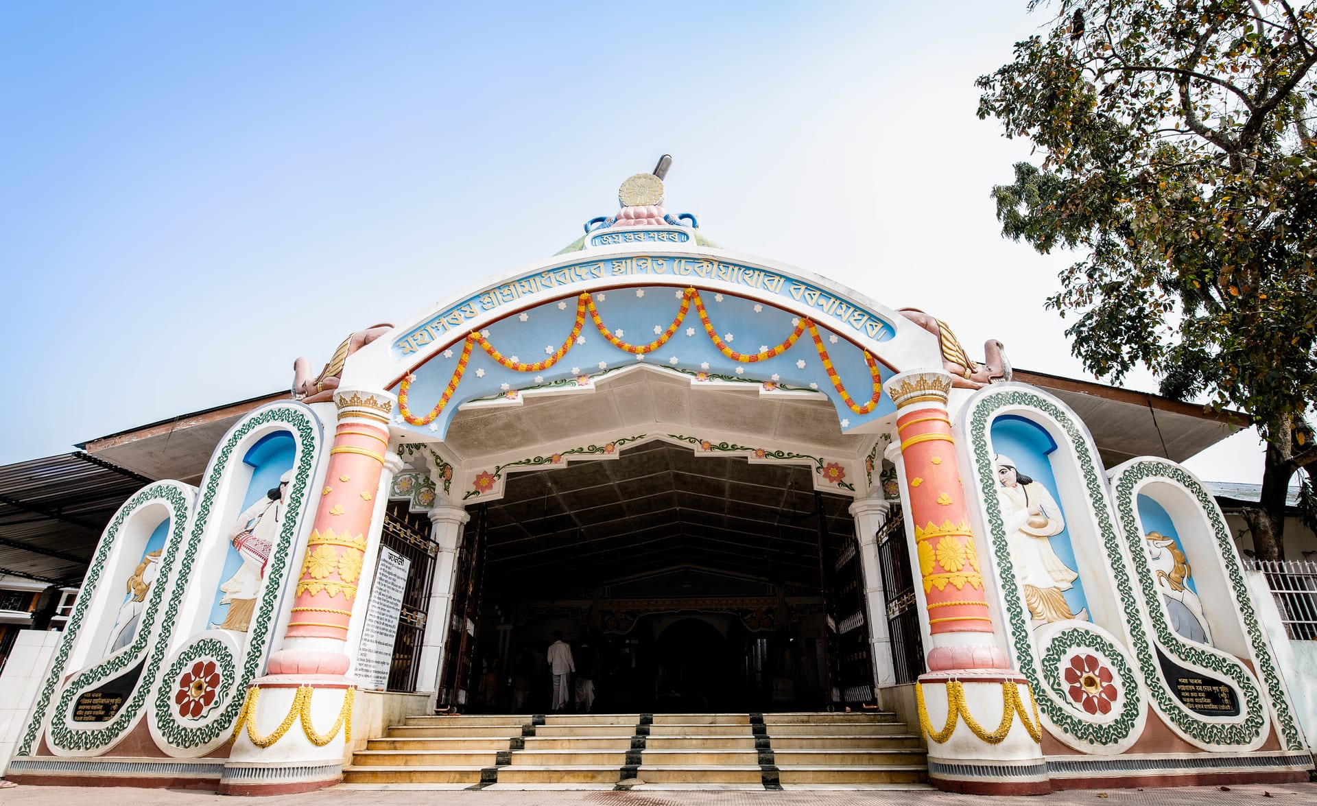 White and pink ornamented entrance porch of the Dhekiakhowa prayer-hall, hung with marigold garlands, with steps leading up to the dark interior