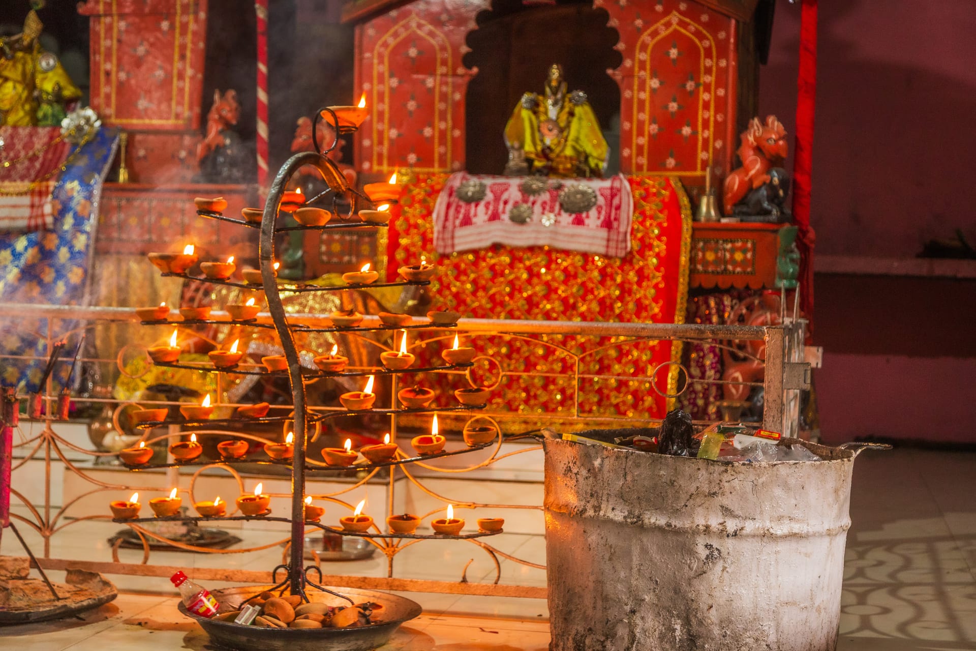 A tiered brass ceremonial lamp glowing with rows of lit wicks before the ornate red-and-gold altar of the namghar at Auniati Sattra