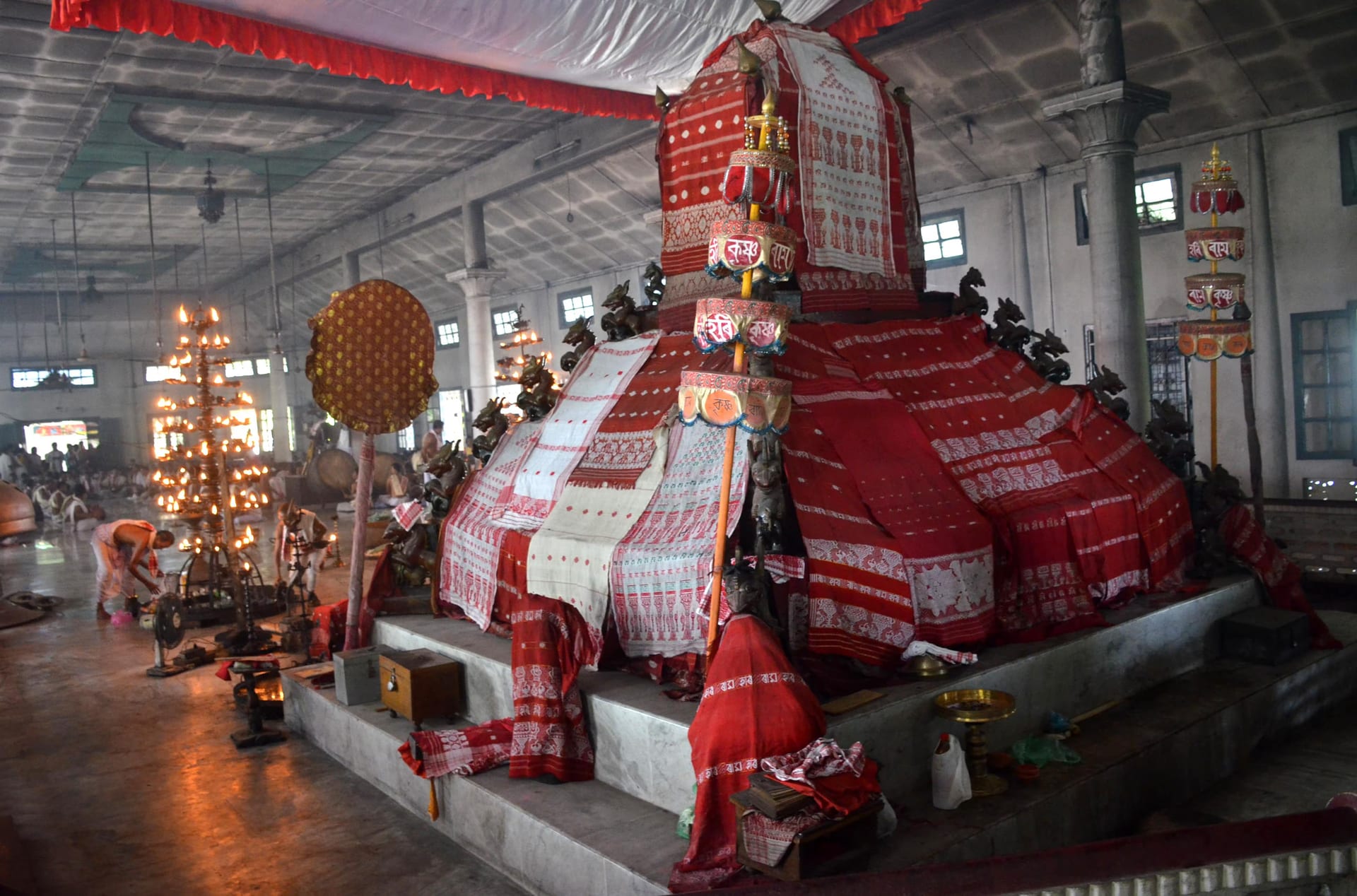 A tall tiered throne inside a pillared prayer hall, draped entirely in offered red and white gamosa cloth, carved winged lions at its tiers and brass lamp-trees alight beside it