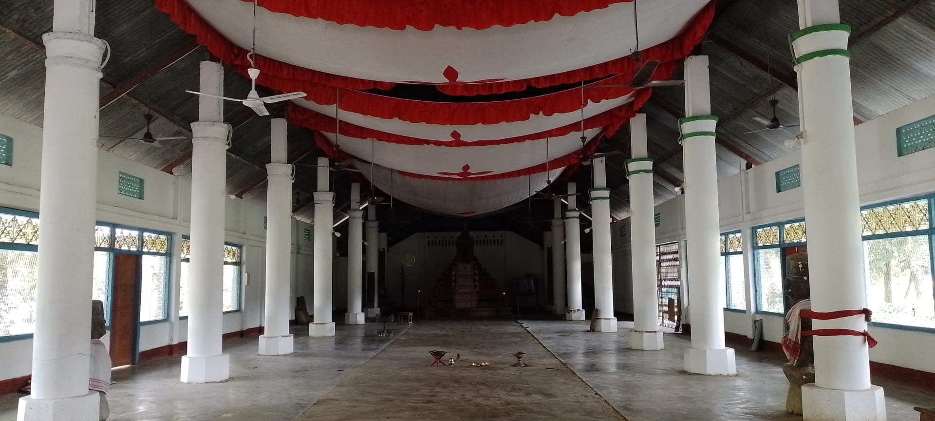 The long pillared prayer hall of the namghar at Bengenaati Sattra, with white columns, a red-and-white ceiling canopy, and the manikut shrine at the far end