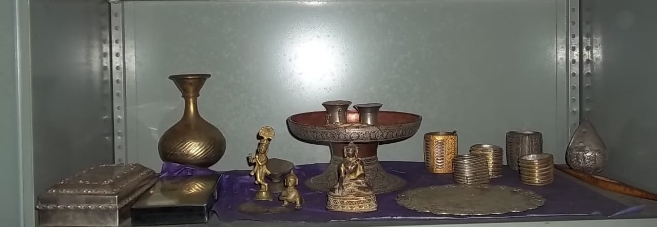 A shelf of gold and silver objects — a gilt Krishna figure, an ornate offering stand, silver caskets and bangles — at Bengenaati Sattra