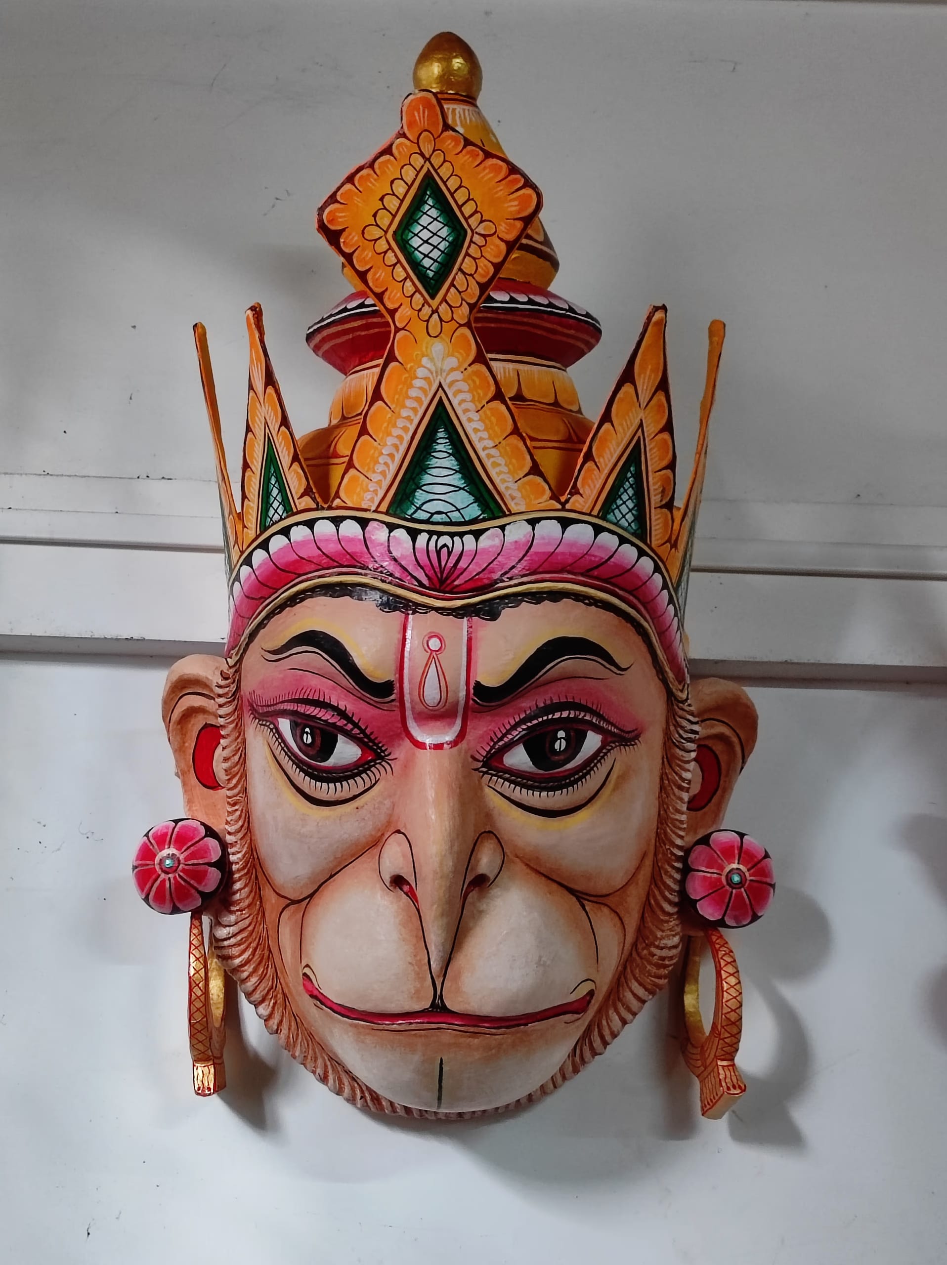 A brightly painted papier-mache mask of Hanuman with a crowned headdress, displayed against a plain white wall