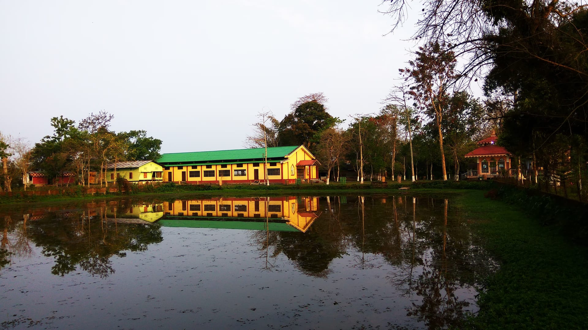 A long pitched-roof sattra building with green roof reflected in a still pond at golden hour, framed by trees, no people