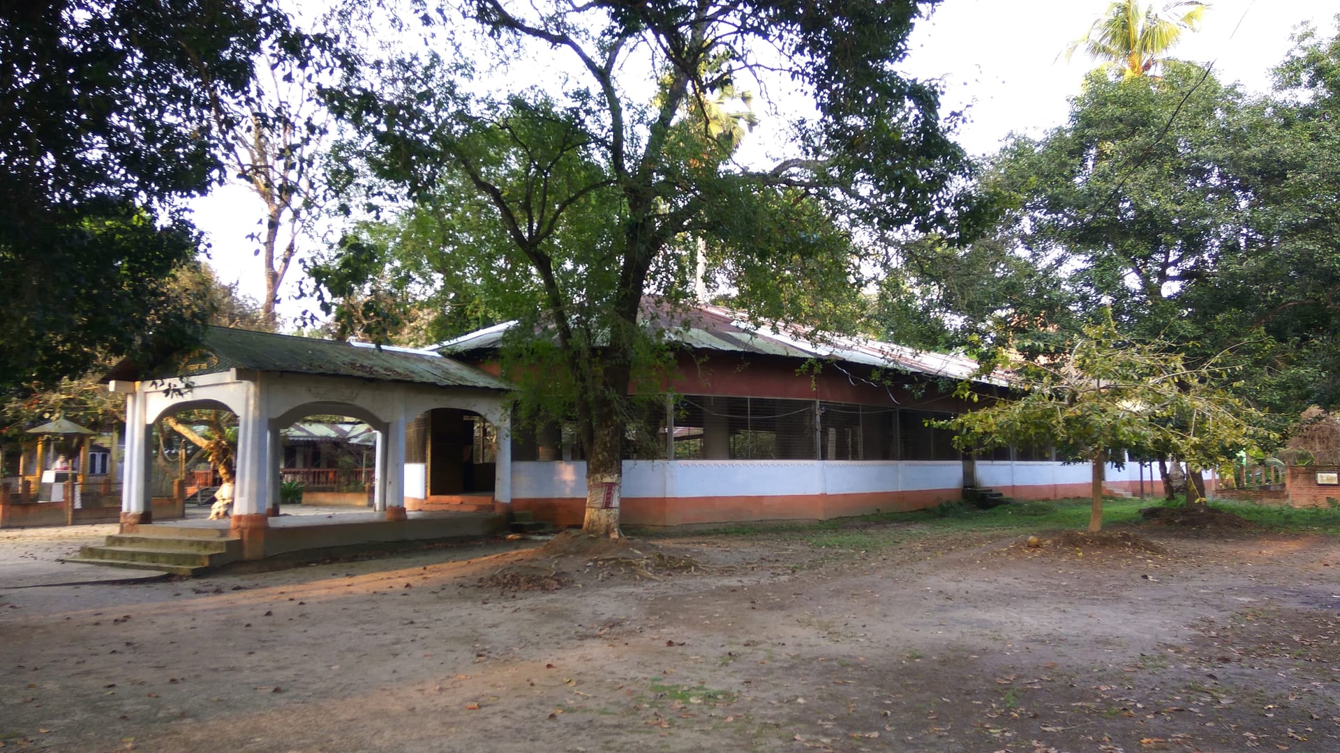 The low tin-roofed prayer hall of Garamur Sattra set among tall trees across a swept earthen courtyard on Majuli