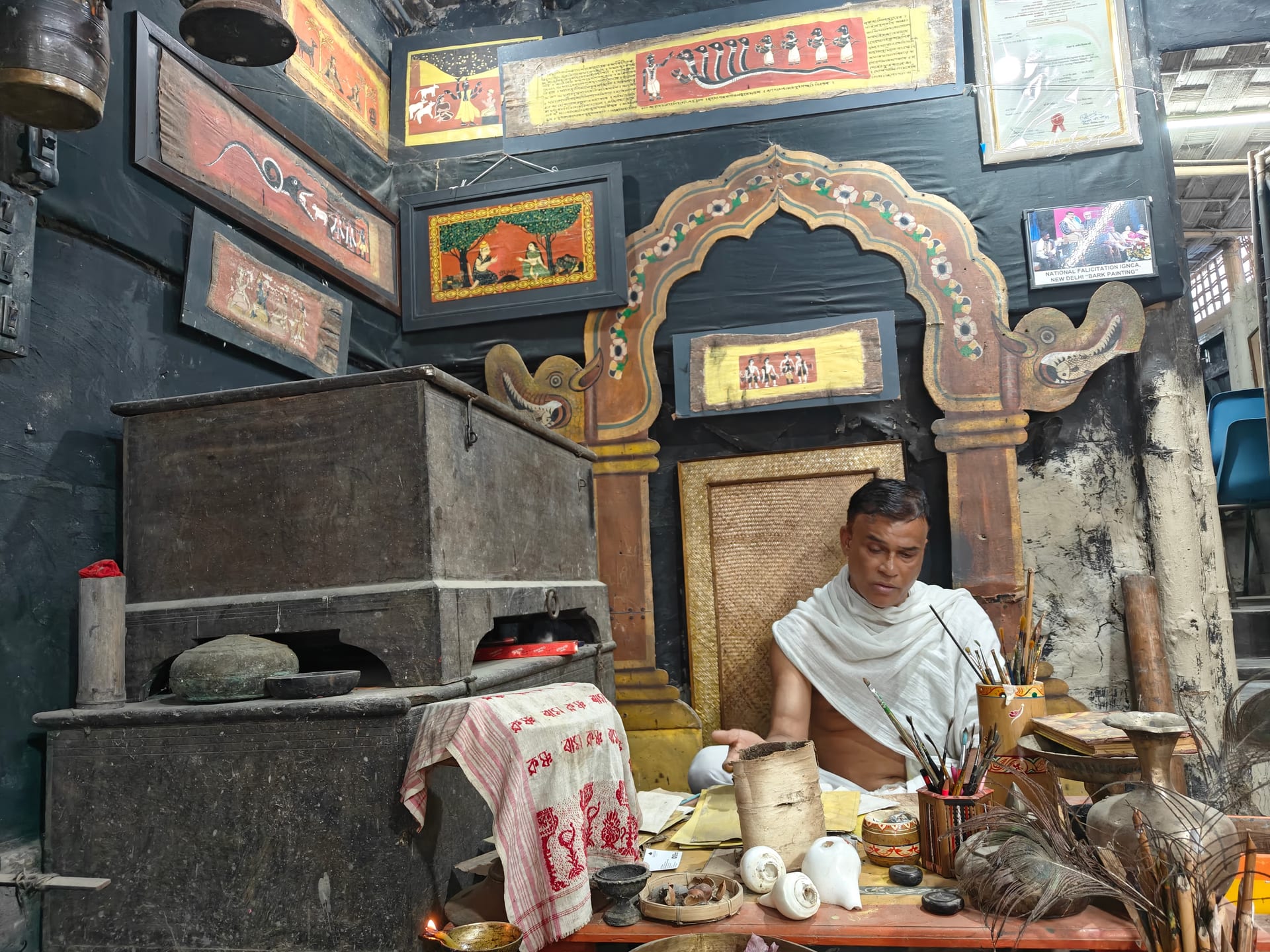 A sattra artist seated beneath a carved and gilded niche hung with painted manuscript folios, working with traditional materials at Garamur on Majuli