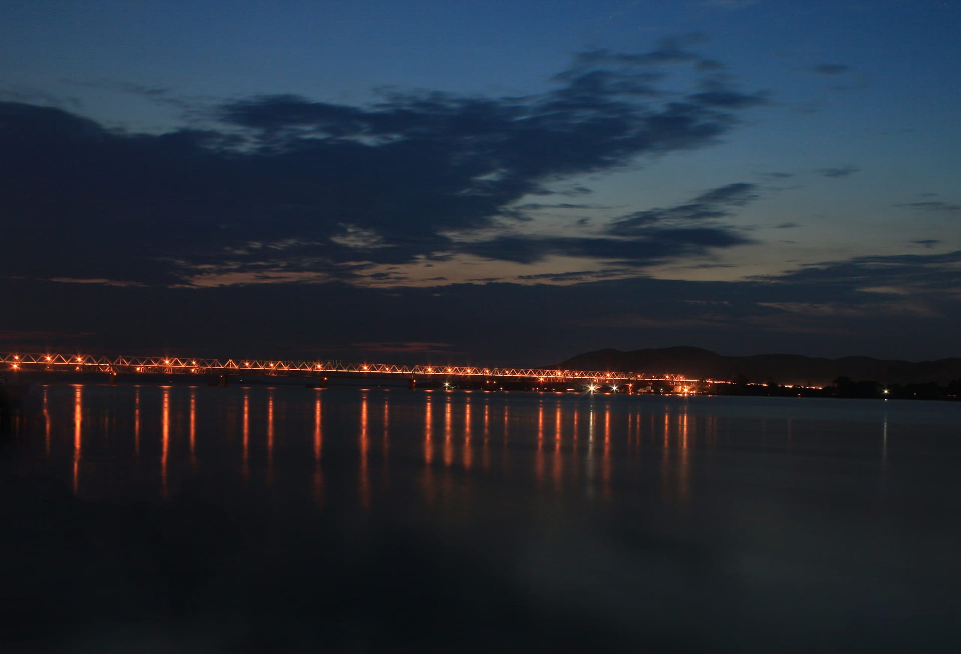 The Saraighat rail-and-road bridge stretched across the Brahmaputra at blue hour, its long line of orange lights mirrored in the still dark water beneath a deep-blue sky streaked with cloud, low hills on the far bank