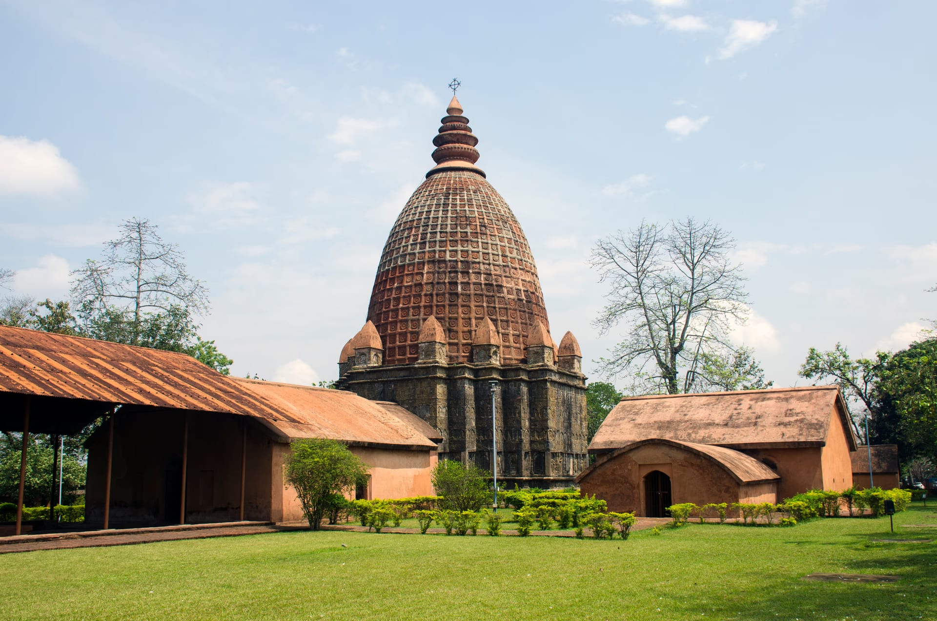 Close three-quarter view of the brick Joydol at Joysagar: a tall ribbed curvilinear Ahom dol tower crowned by a tiered amalaka and trident finial, rising over a square sanctum, with a low pillared hall to the left and a barrel-roofed brick shrine to the right under a blue sky