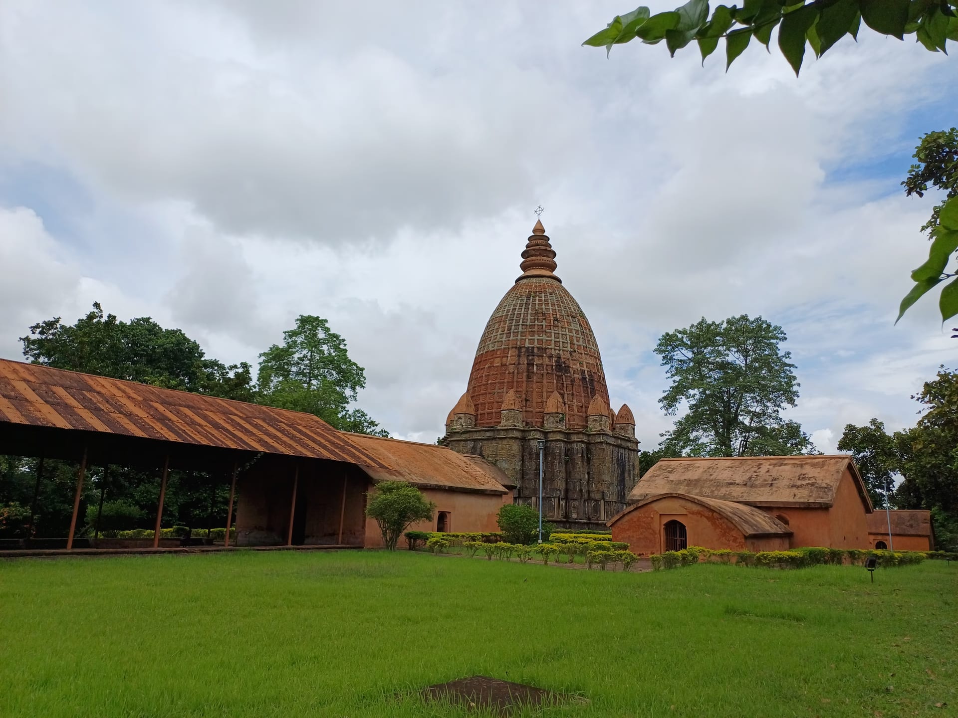 Low-angle view across a green lawn to the brick Joydol at Joysagar: a ribbed curvilinear Ahom dol tower crowned by a tiered amalaka and trident finial over a square carved sanctum, with an open pillared hall to the left and a barrel-roofed brick shrine to the right, under a dramatic monsoon sky