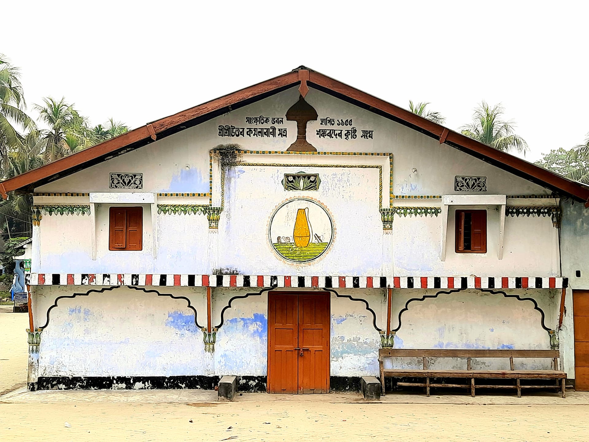 Whitewashed prayer-hall facade of the Uttar Kamalabari Sattra on Majuli, with an Assamese inscription in the gable, a central doorway and a painted emblem