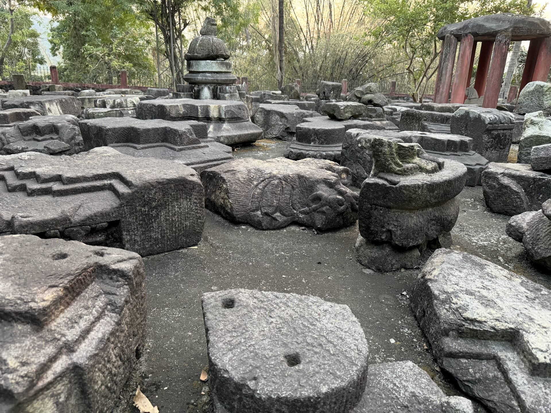 A wide field of scattered carved granite blocks, plinths and a stacked stone finial among trees at Madan Kamdev