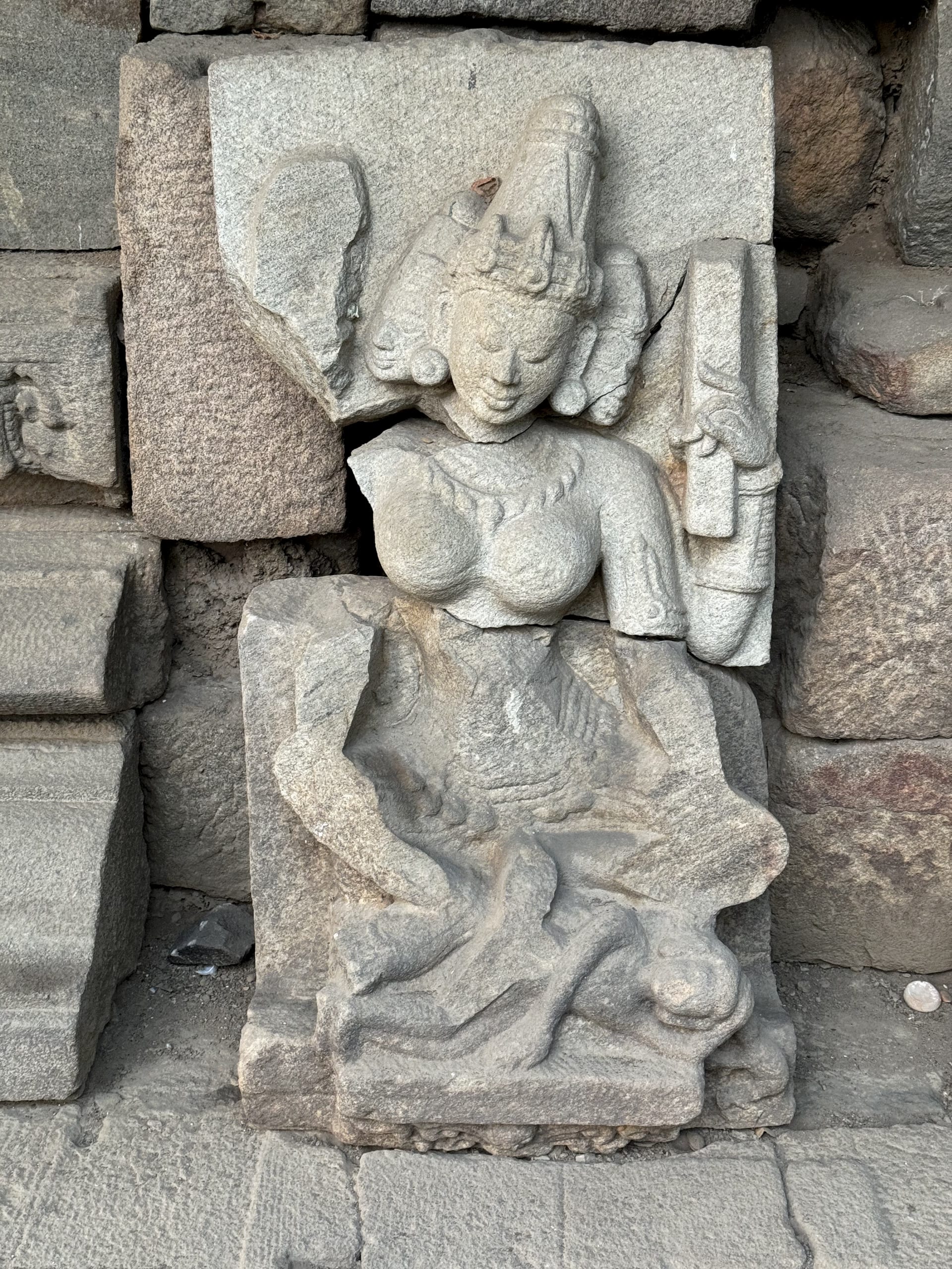 A finely carved crowned female figure in stone with elaborate headdress and jewellery among stacked temple blocks