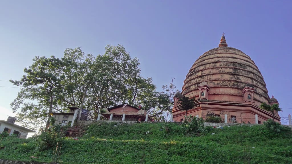 The brick-red ribbed beehive dome of the Navagraha temple rising above the green slope of Chitrasal hill, with smaller shrines beside it