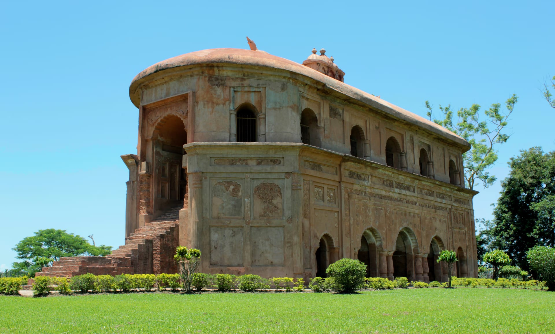 Three-quarter view of the Rang Ghar against a clear blue sky, showing its curved oval end, carved side wall and boat-shaped roof above a green lawn