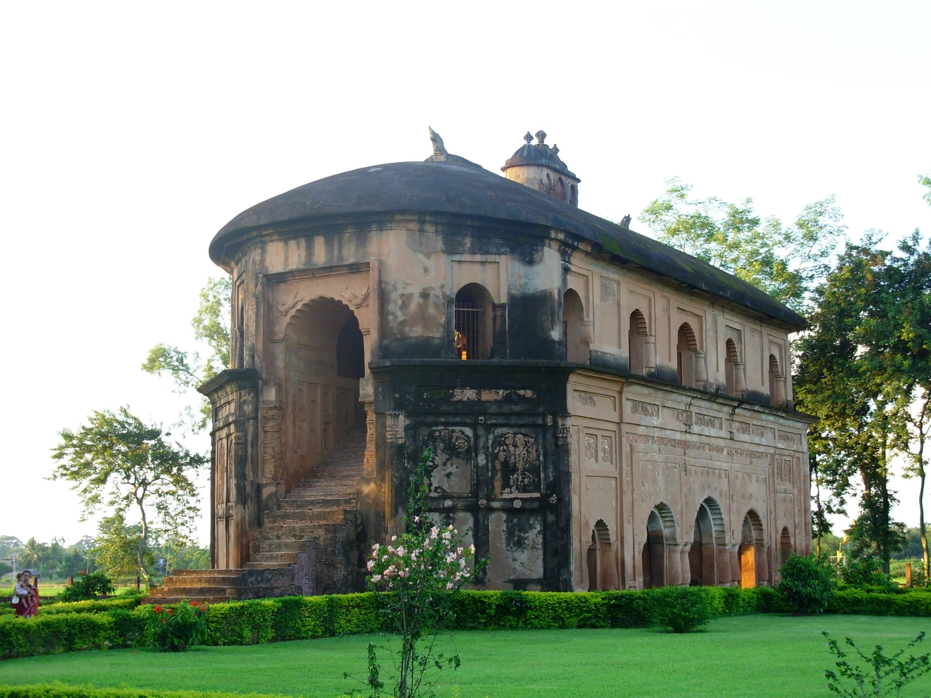 Rear three-quarter view of the Rang Ghar in warm afternoon light, showing the external masonry ramp that climbs to the upper gallery, with carved panels on the curved end and green surroundings