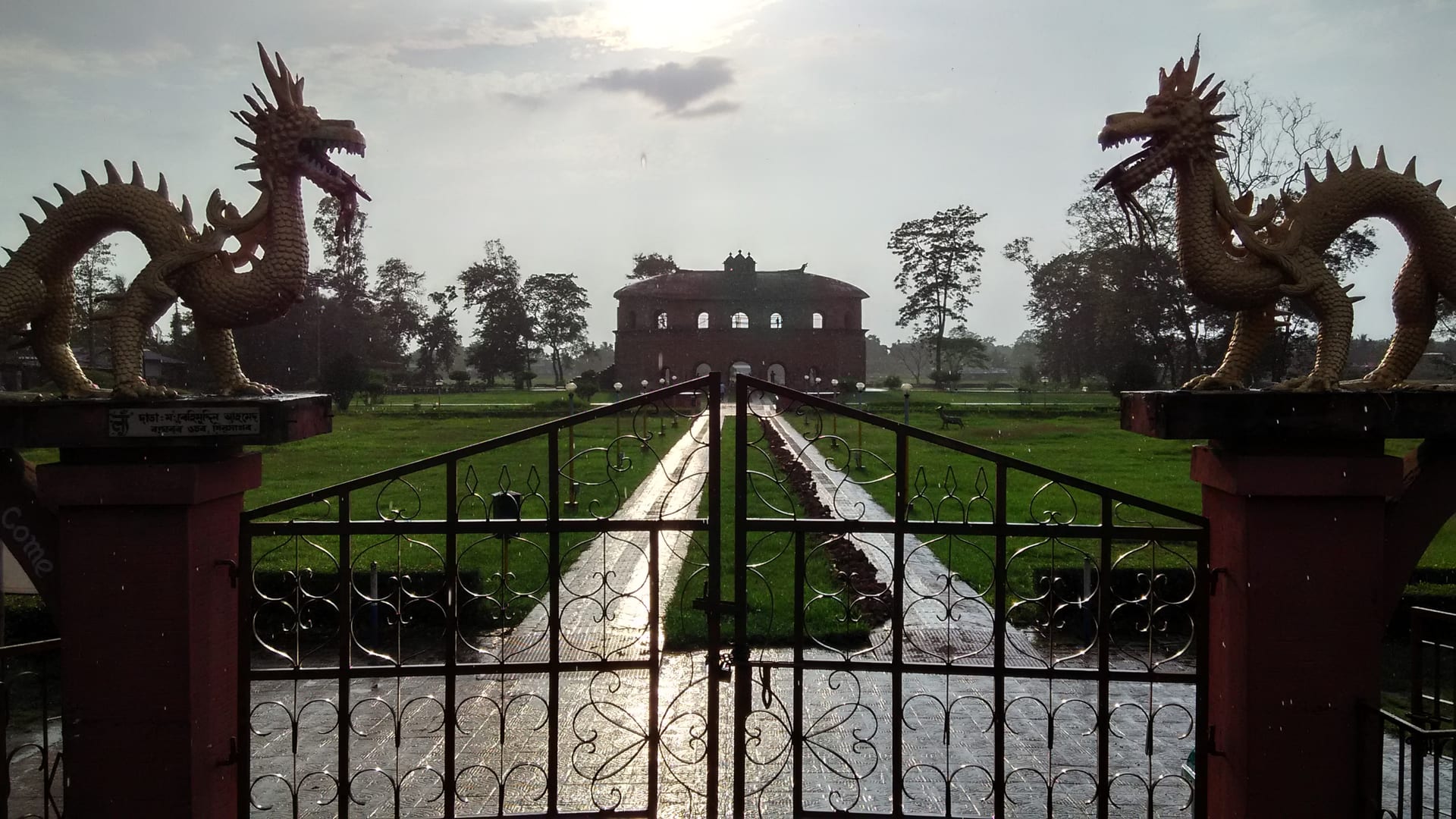 The Rang Ghar seen down a long axial garden, framed by a pair of golden dragon statues mounted on the gateposts above a wrought-iron gate, the oval pavilion centered in the distance