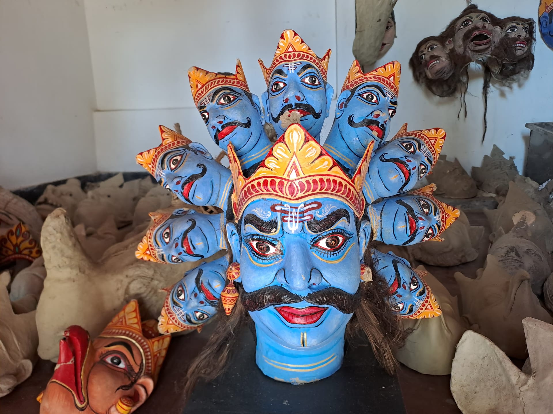 Large blue ten-headed Ravana mukha mask crowned with smaller faces, standing among unpainted clay masks in the Samaguri workshop on Majuli